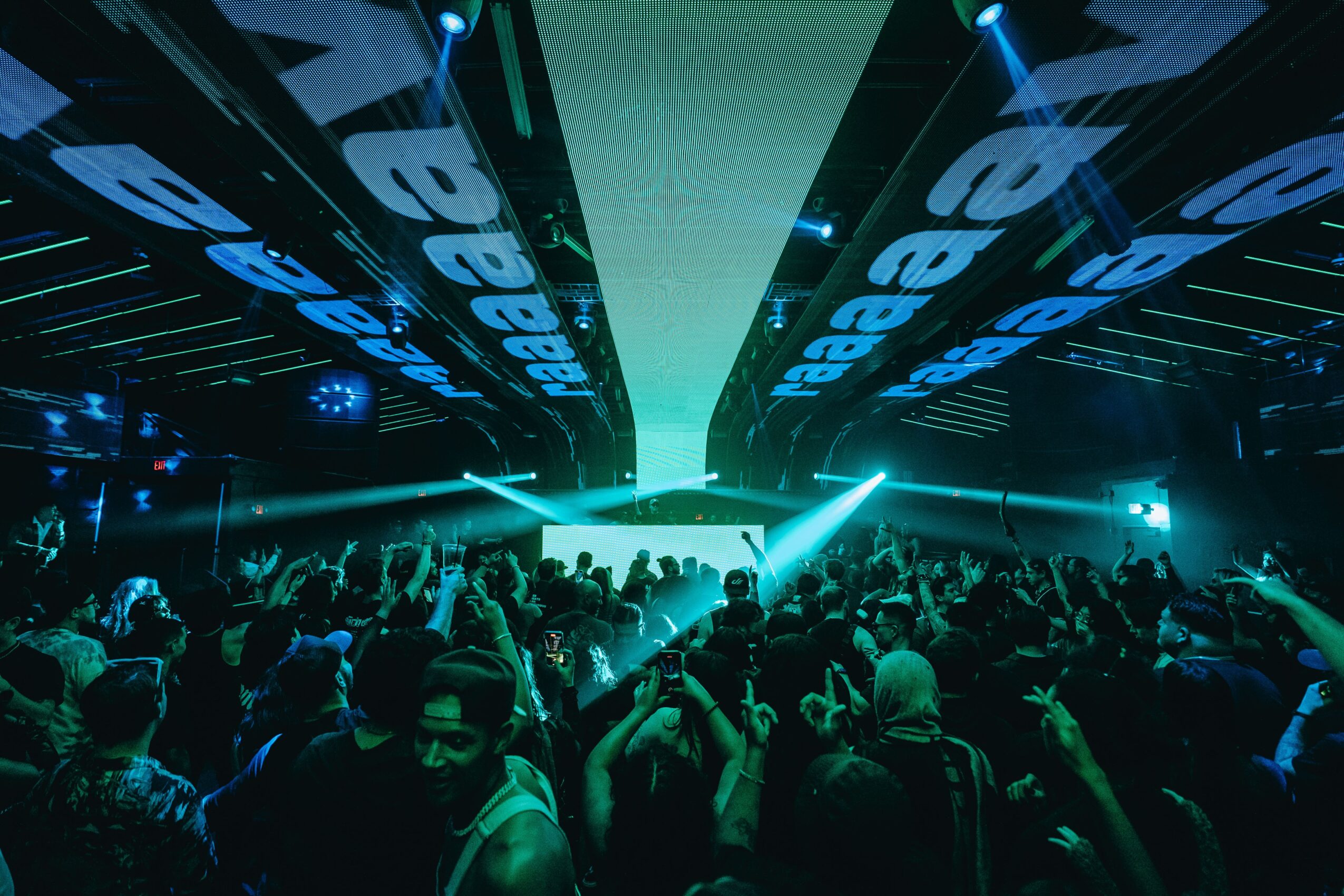 Crowd at a concert or event with bright lights and digital screens overhead, people dancing and enjoying the atmosphere.