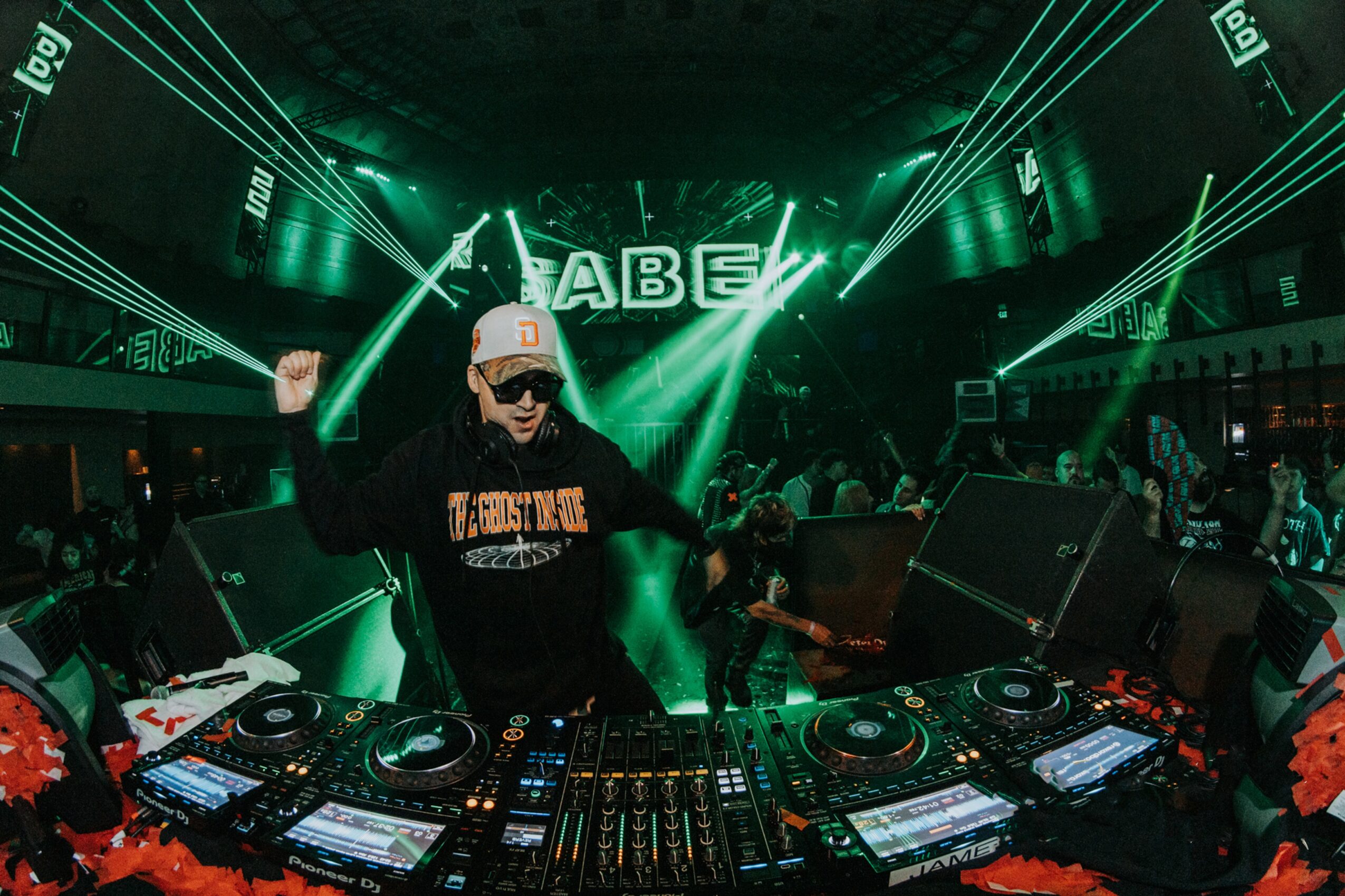 DJ wearing sunglasses and a cap, performing with green laser lights in a club. DJ equipment visible in foreground.