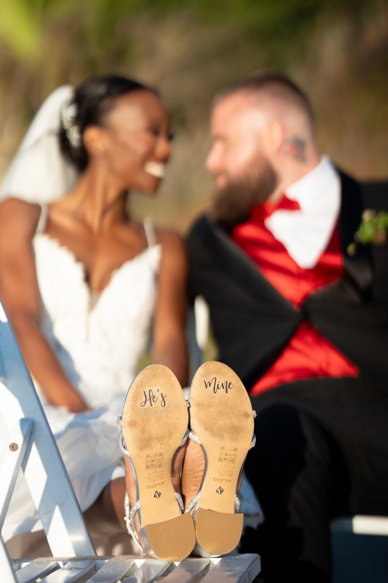 Bride and groom smiling at each other outdoors, with sandals in foreground, wedding attire, and blurred background.