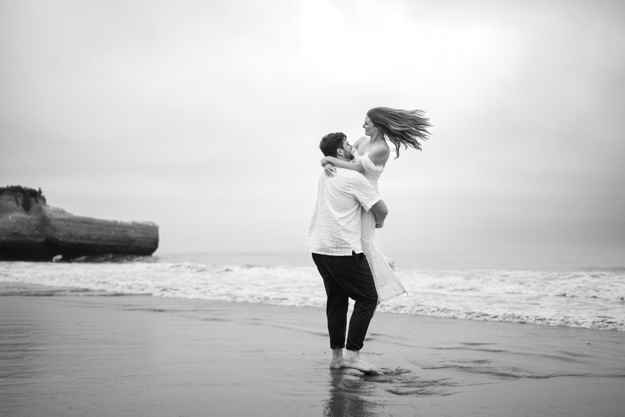 A couple on the beach, the man lifting the woman, her hair flowing, ocean and cloudy sky in background.