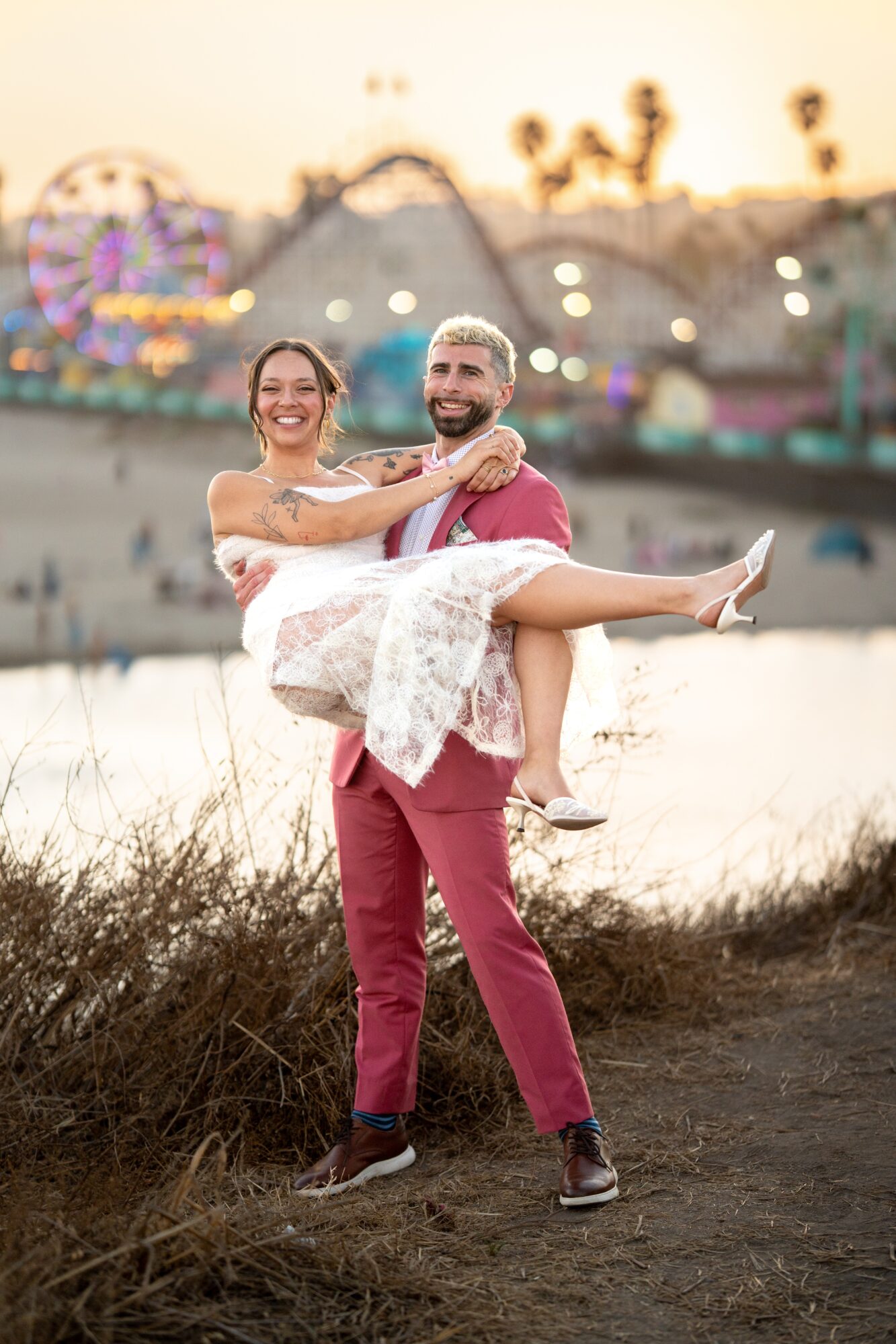 Man in pink suit carrying smiling woman in white dress outdoors near water, with blurred amusement park in background.