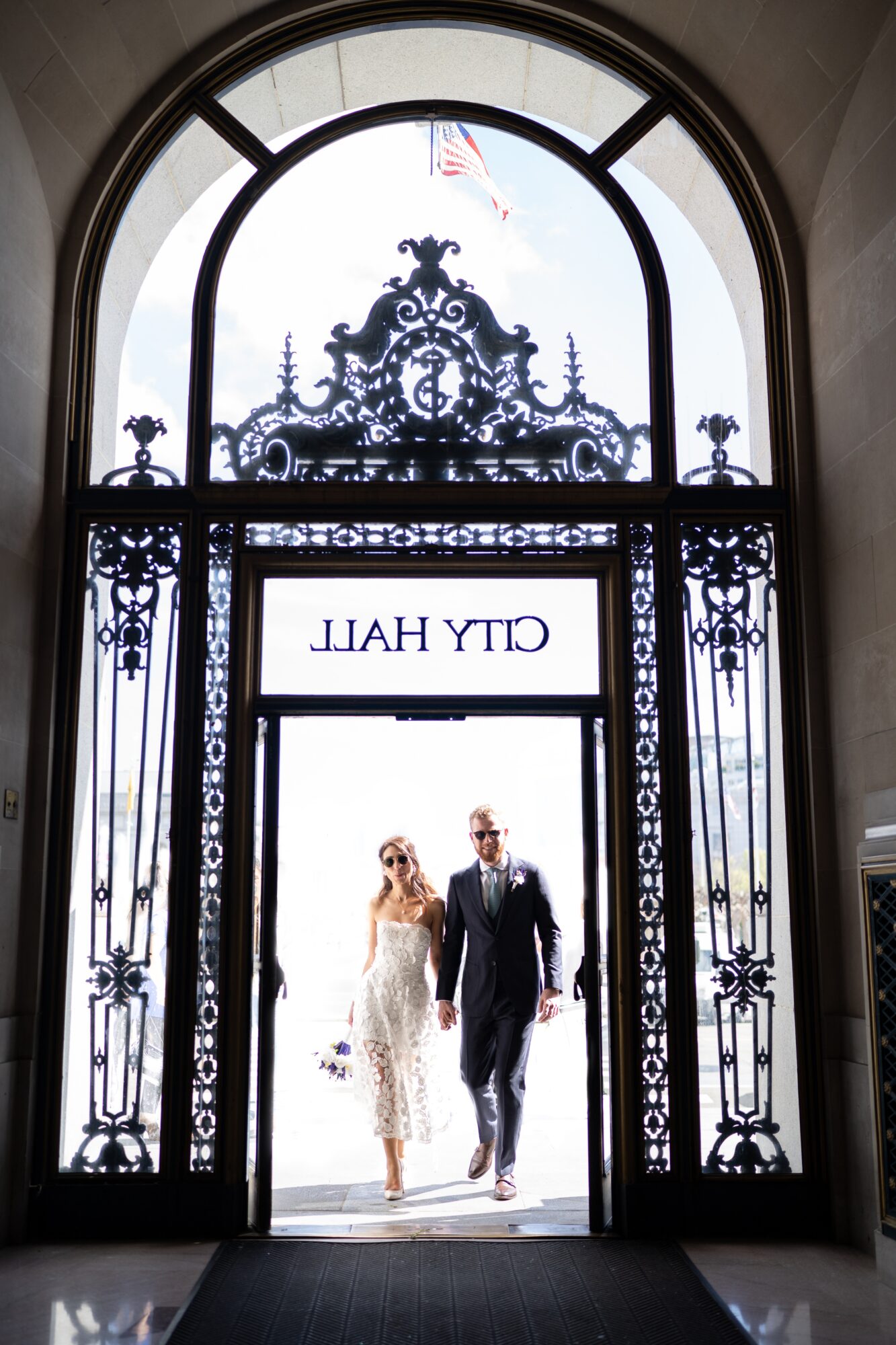 Couple walking through ornate doorway with decorative ironwork and large arched window above, city hall sign visible, American flag in background.