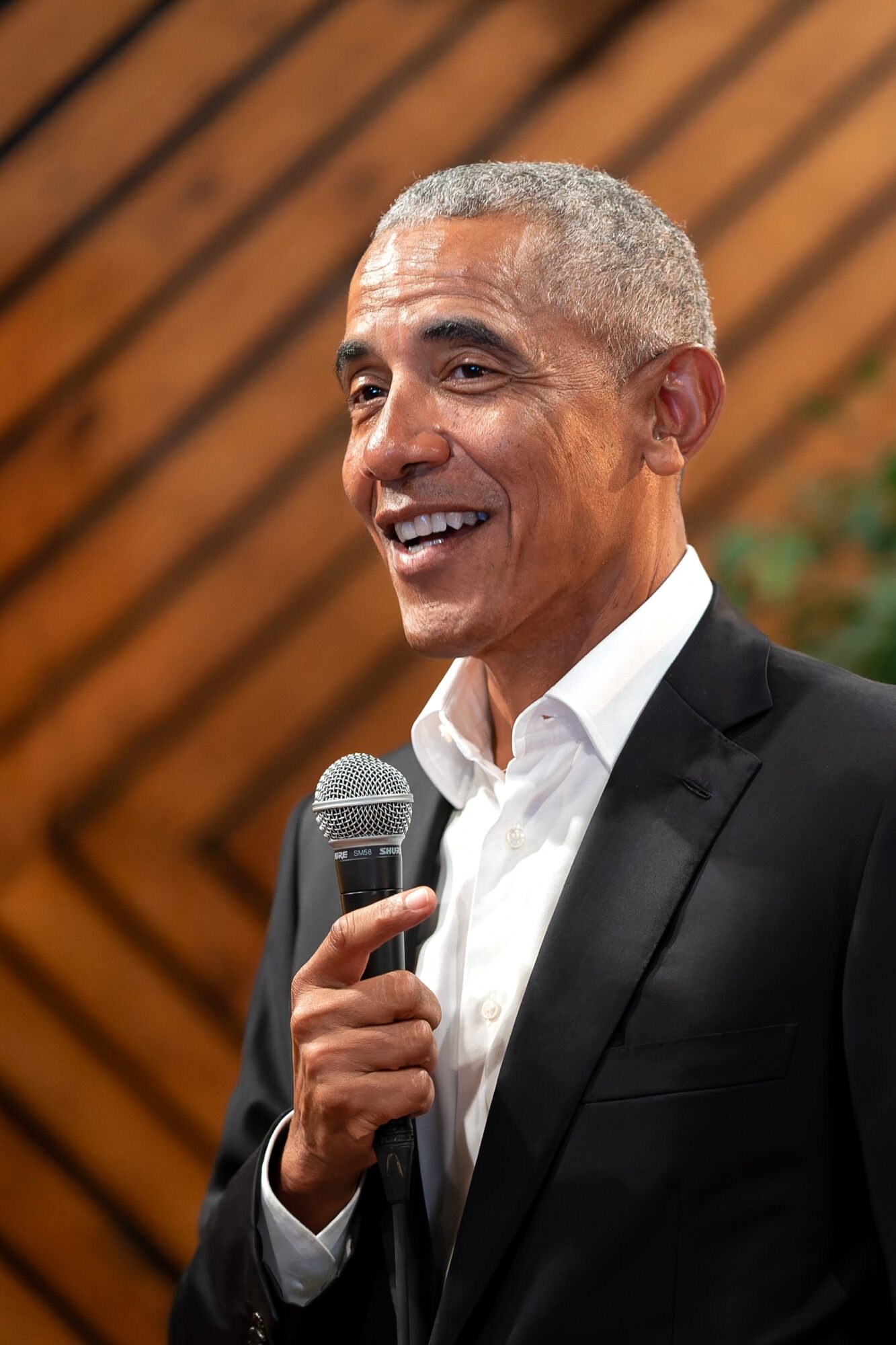 Man in suit speaking into a microphone, smiling, with a wooden background.