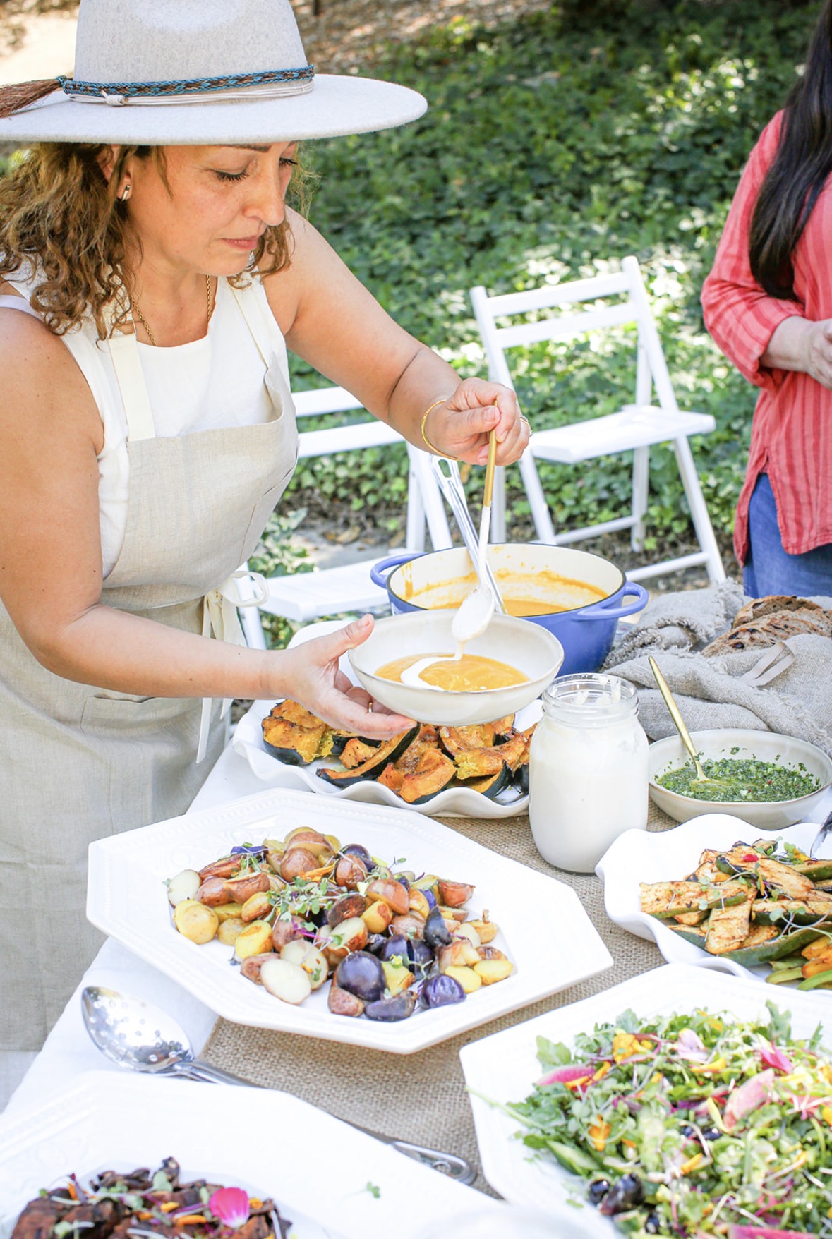 Woman in a hat serving soup at outdoor gathering with food and greenery in background.