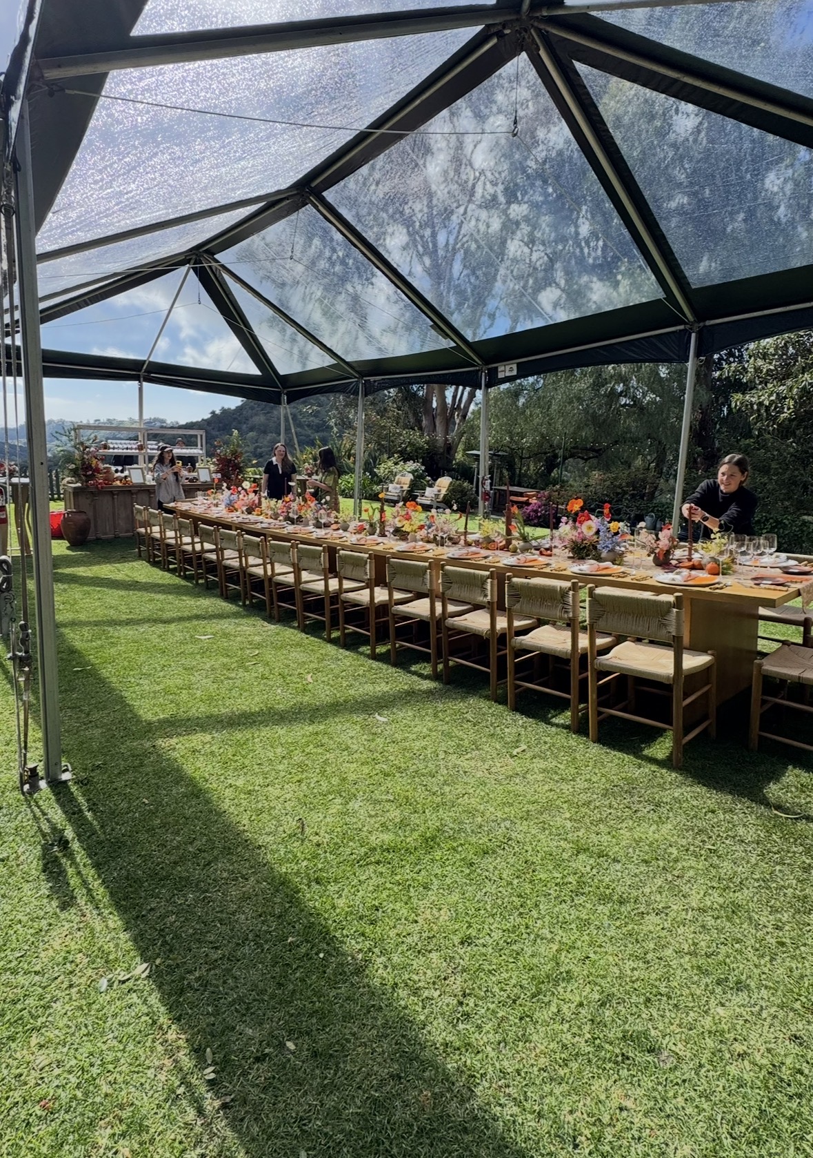 Long outdoor table set under a glass tent with chairs, flowers, and people, on a grassy area with trees and sky.