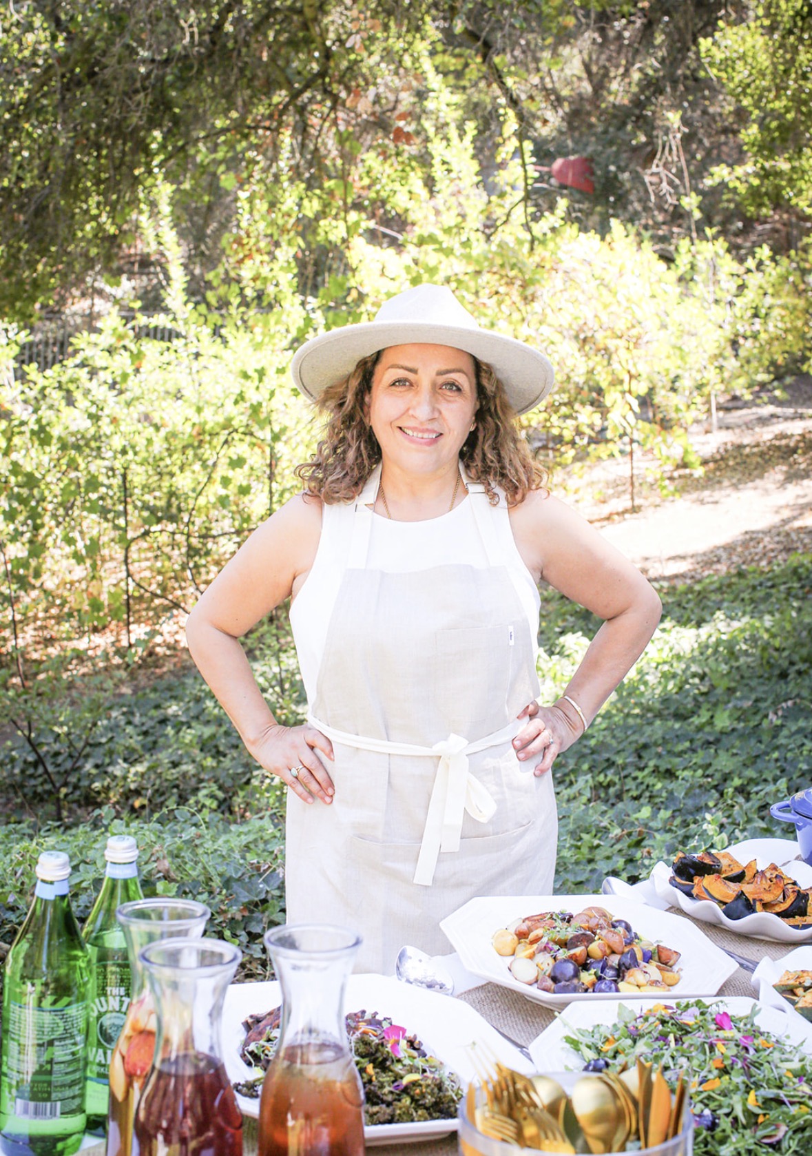 Woman in a white dress and hat standing outdoors with food and drinks on table, smiling, surrounded by trees and greenery.