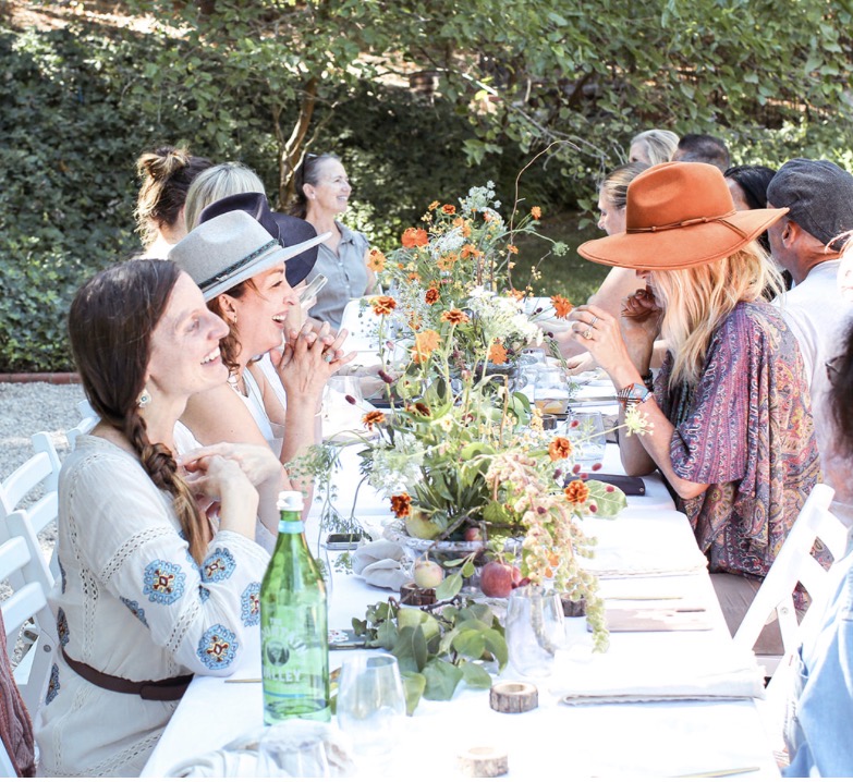 People sitting at a table outdoors with flowers, drinking and talking, under trees, during daytime.