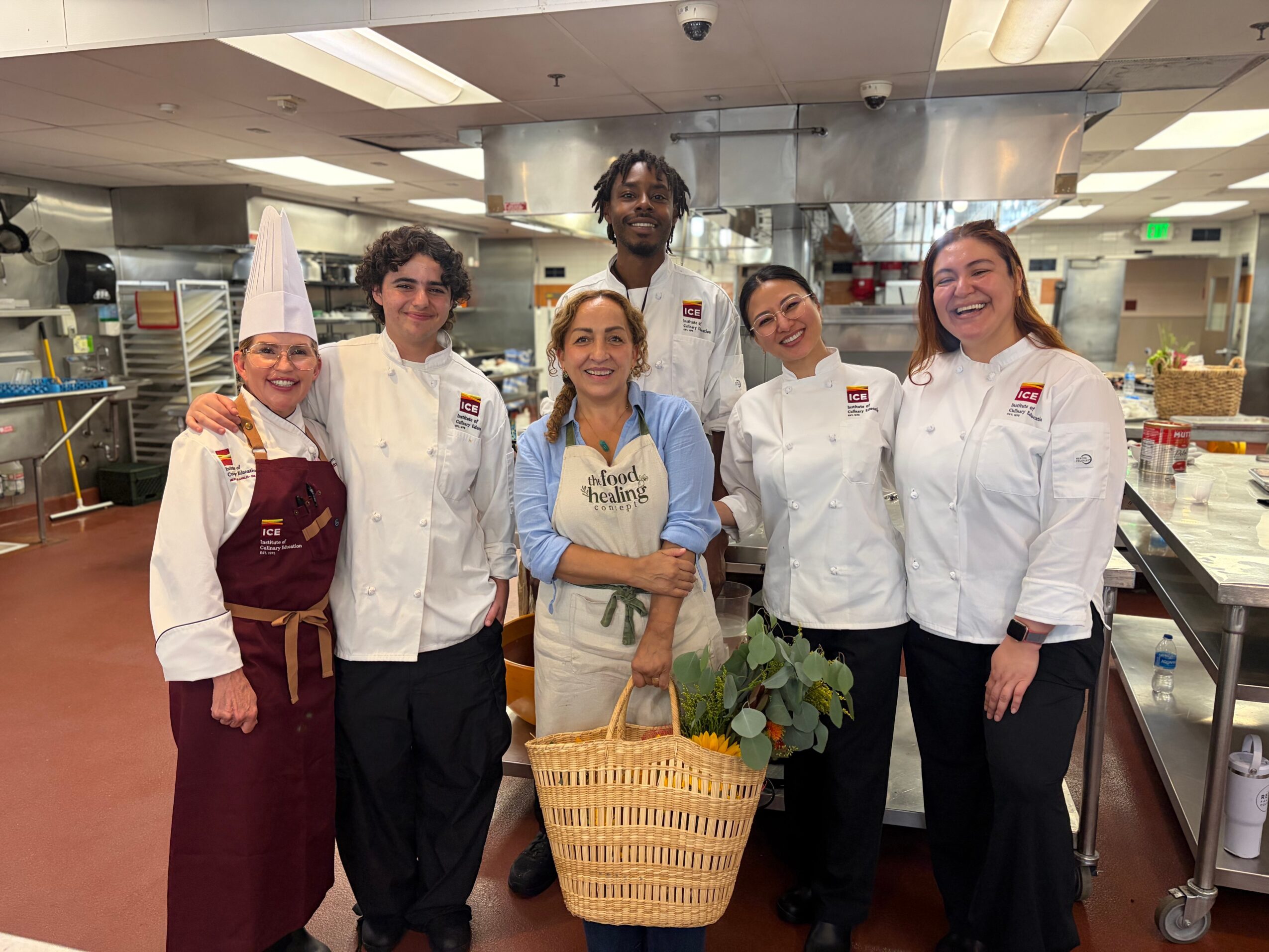Group of six chefs and a woman holding a basket with plants in a commercial kitchen.