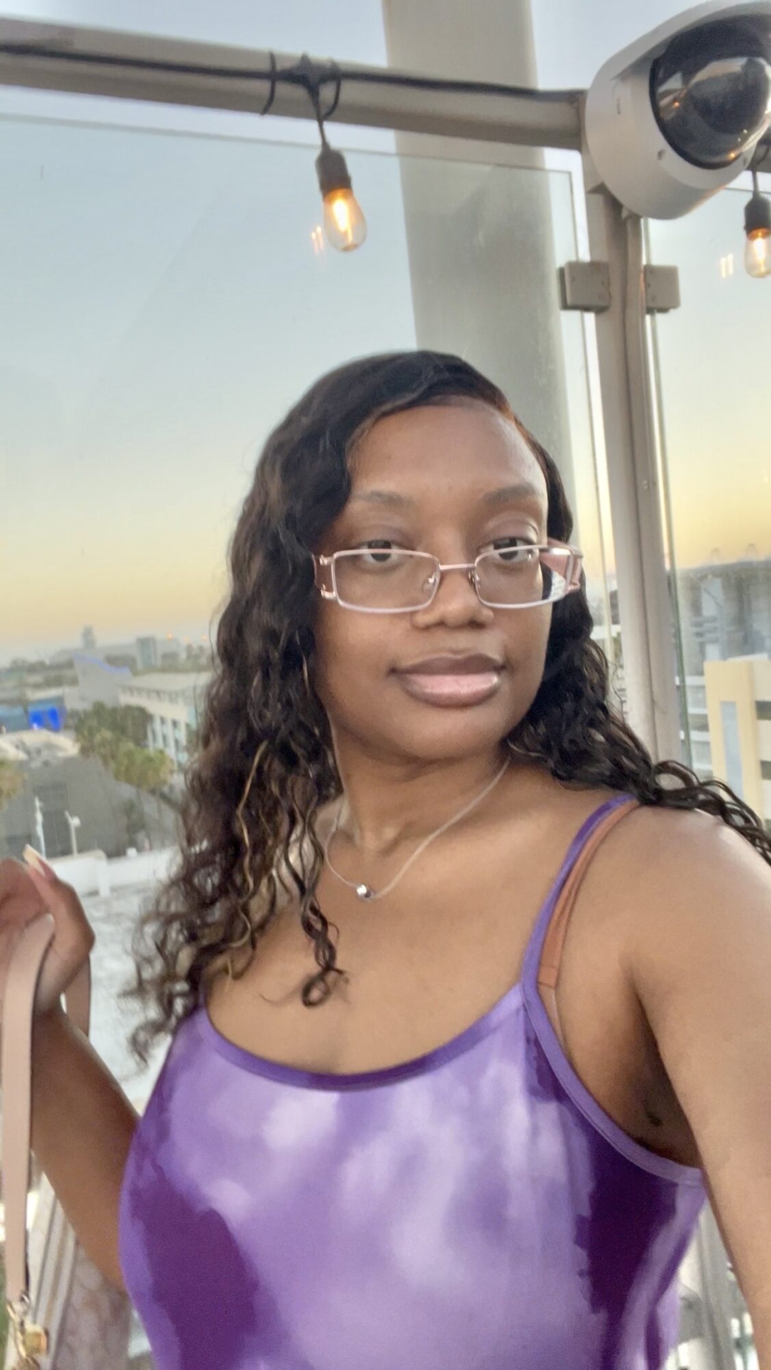 Woman with glasses and wavy hair wearing a purple satin top, standing outdoors with cityscape background.