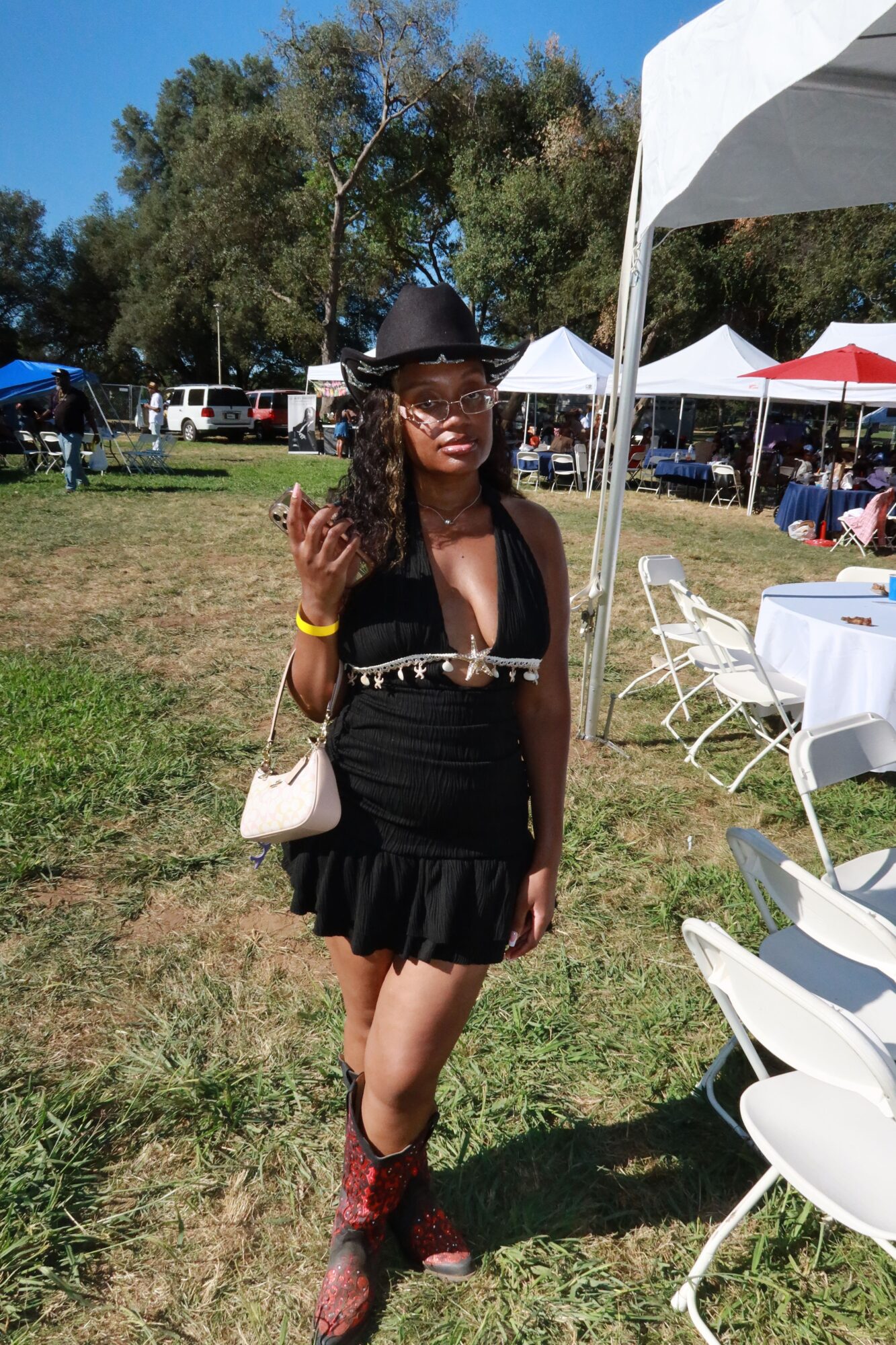 Woman in black dress and sunglasses standing outdoors, waving, with chairs and tables nearby.