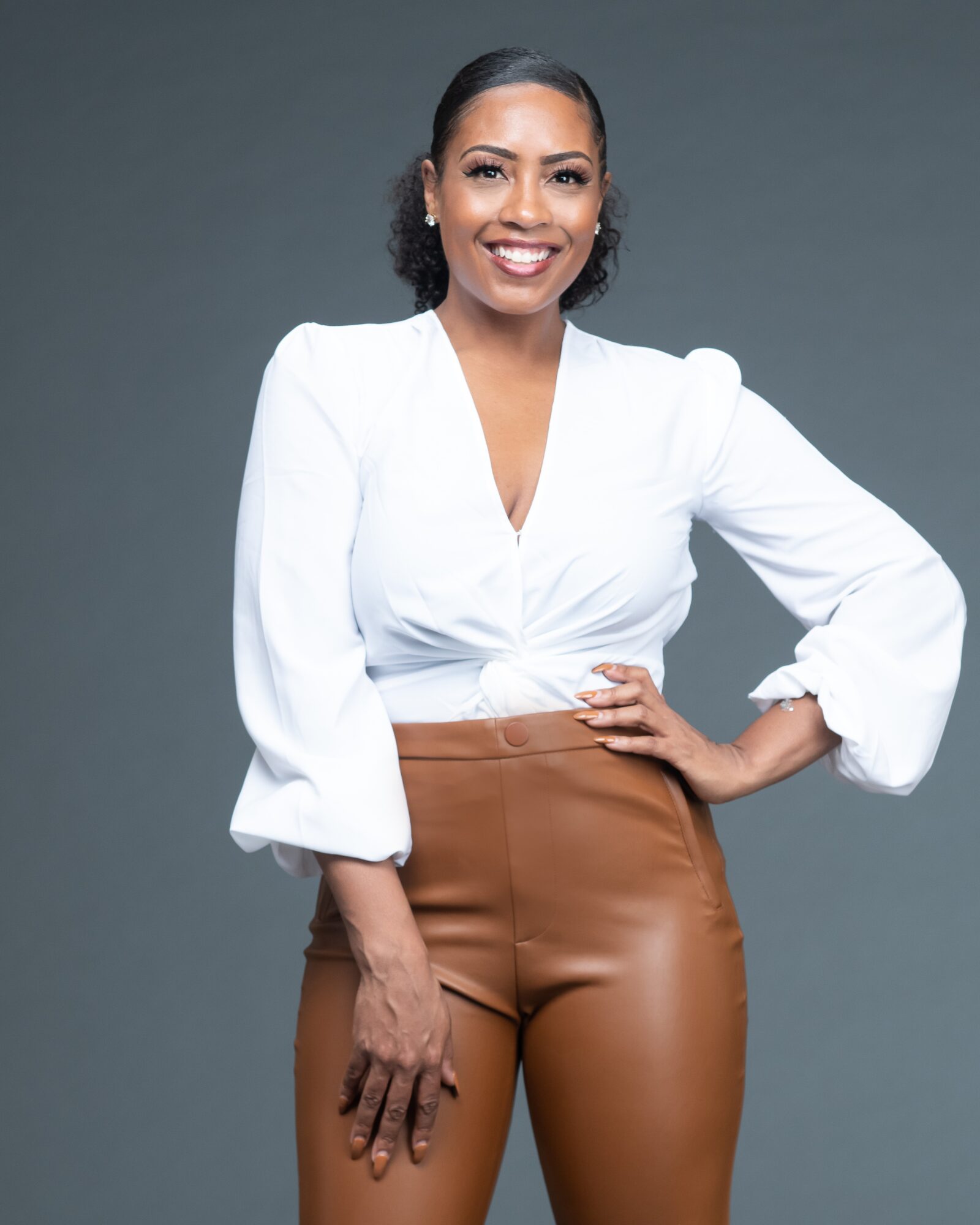 Woman smiling, wearing a white blouse and brown pants, standing with one hand on her hip against a gray background.