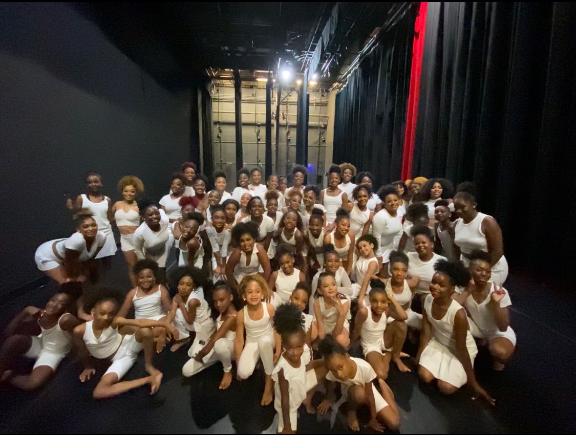 Group of children and adults in dance costumes on stage, smiling and posing for a photo.