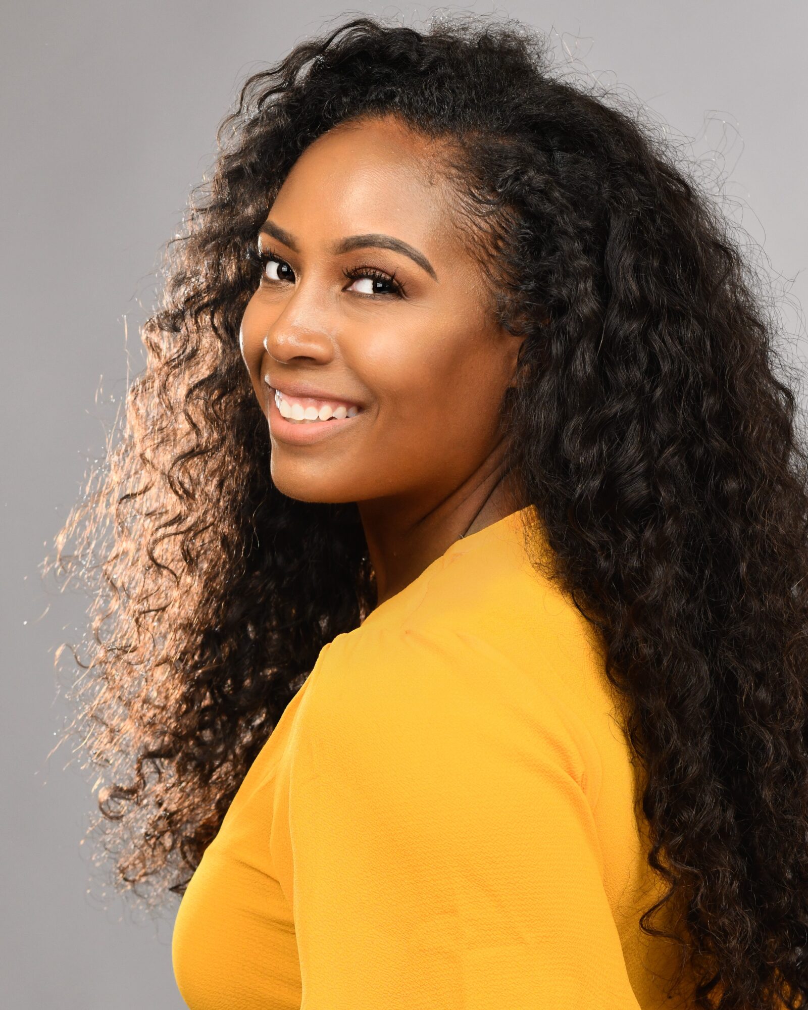 Smiling woman with curly hair wearing a yellow top, facing slightly to the right, against a gray background.