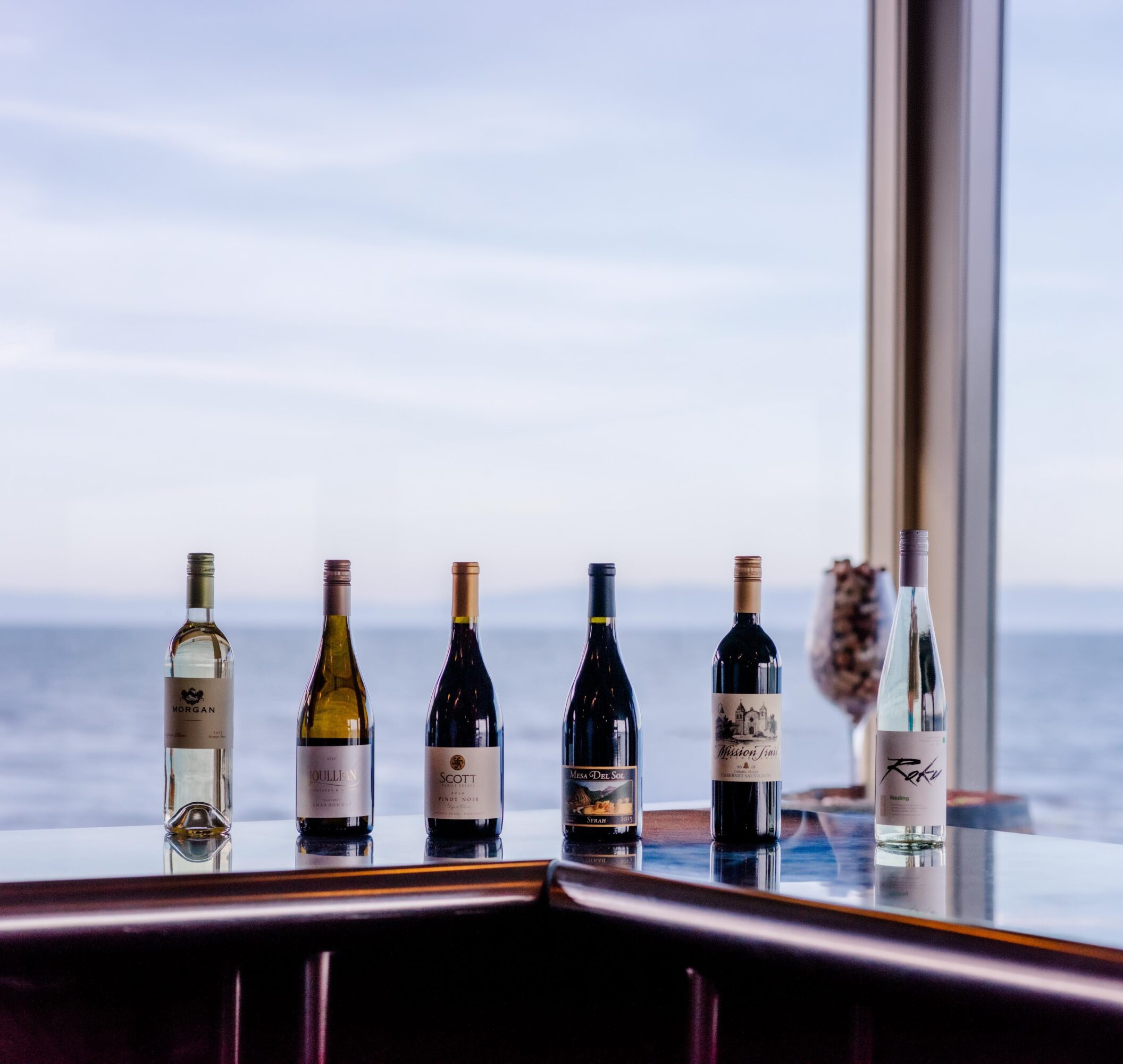 Six bottles of wine and a glass vase with flowers on a table near a window with a view of the ocean.