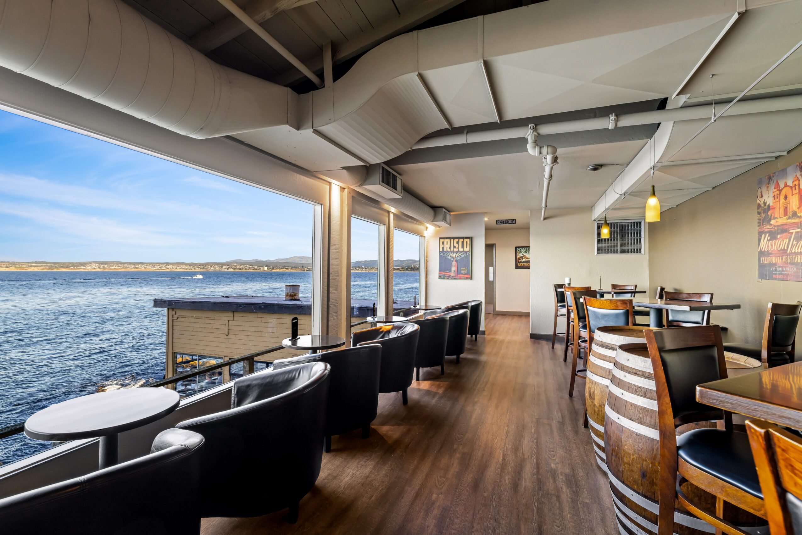Interior of a restaurant with large windows overlooking water, black chairs, and wooden tables, with ceiling pipes visible.