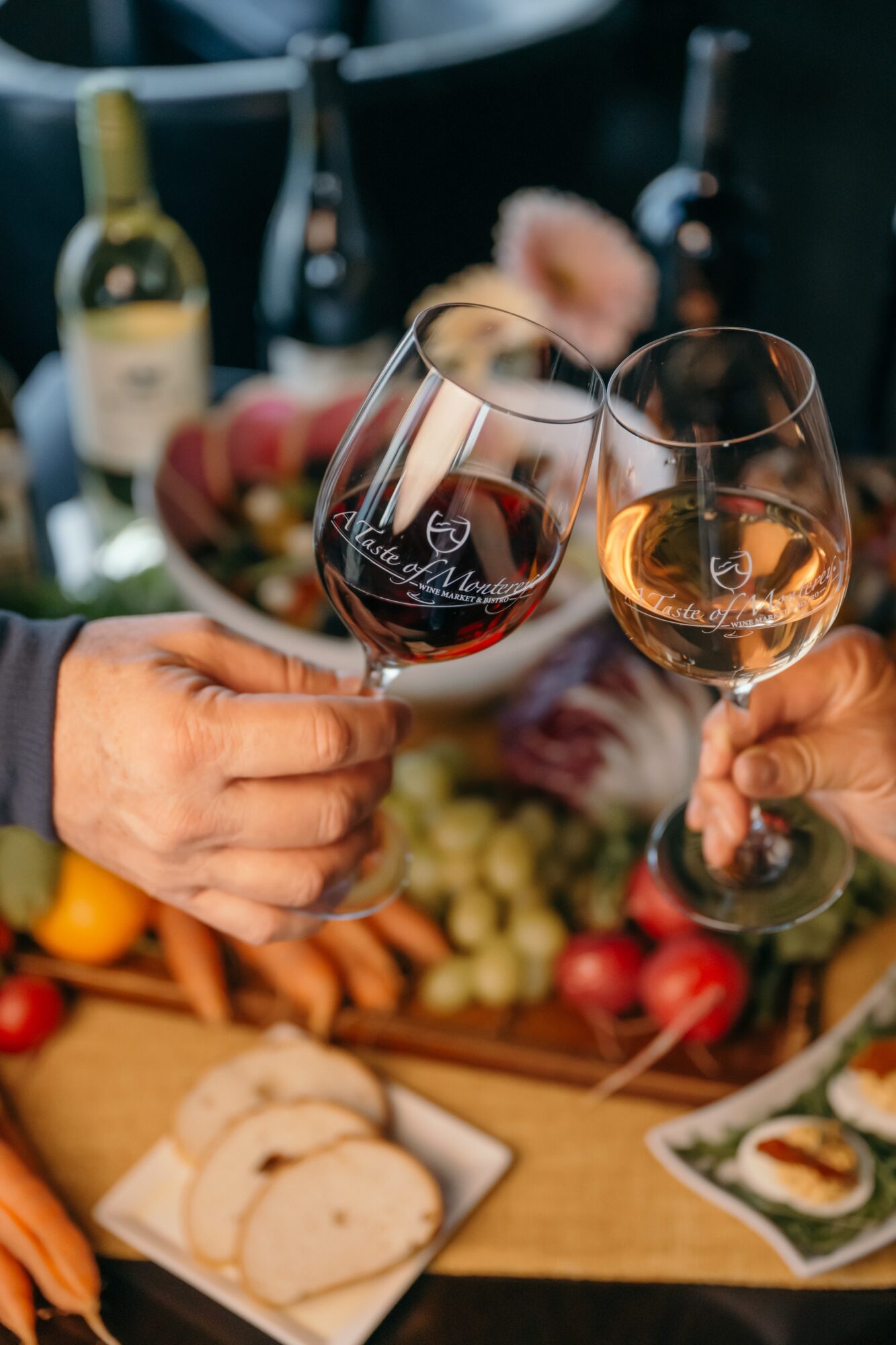 Two people clink glasses of red and white wine over a table with food and drinks, blurred background.
