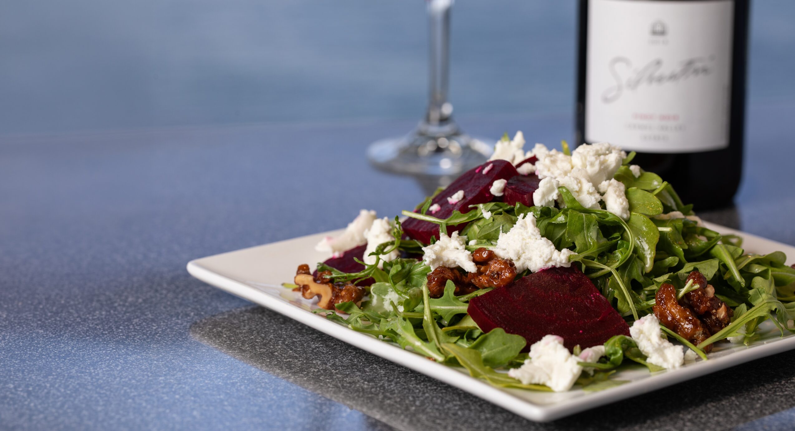 Fresh salad with greens, beets, and cheese on a white plate, with a wine bottle and glass in the background.
