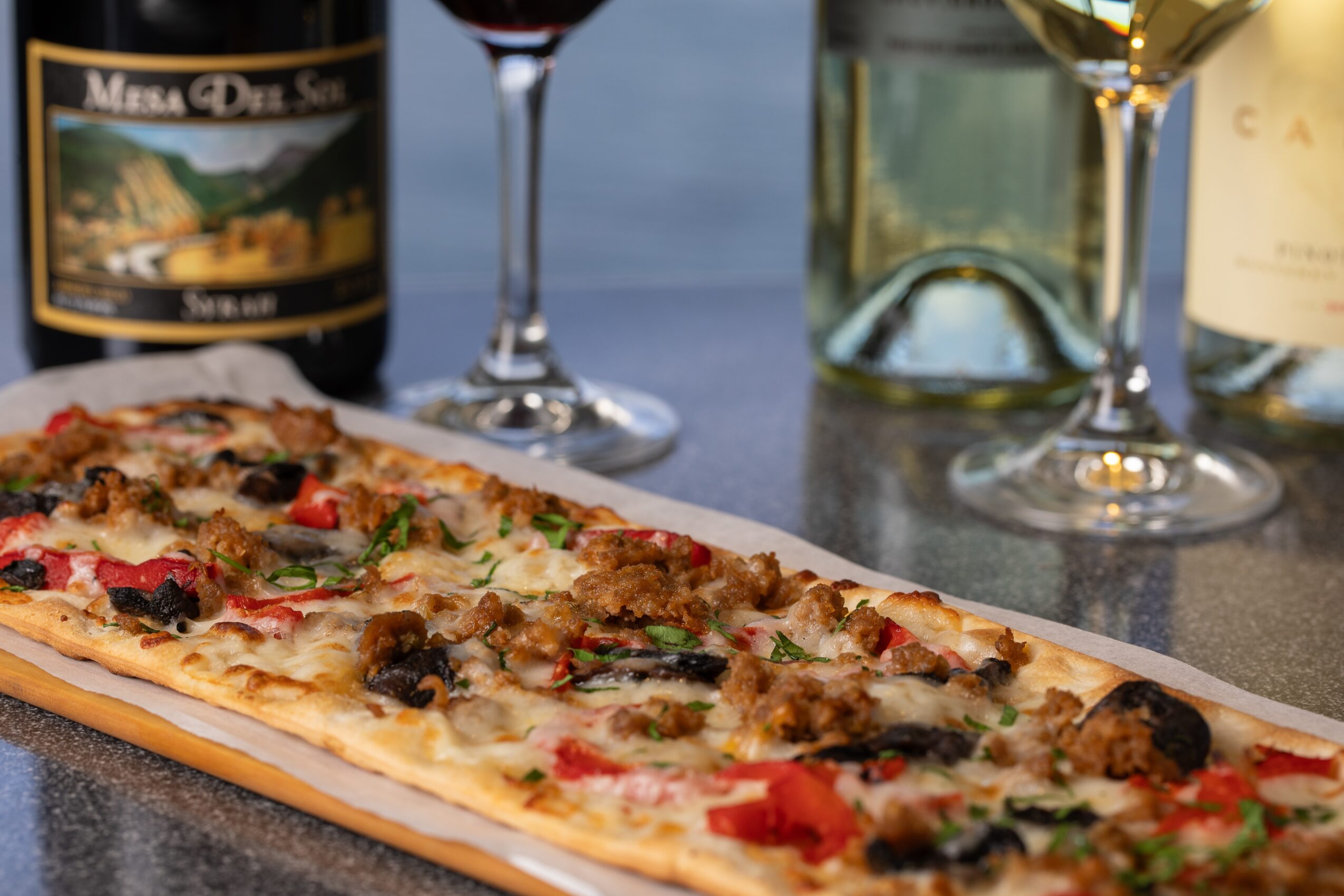 Long rectangular pizza with toppings including vegetables and meat, on a table with wine glasses and bottles in the background.