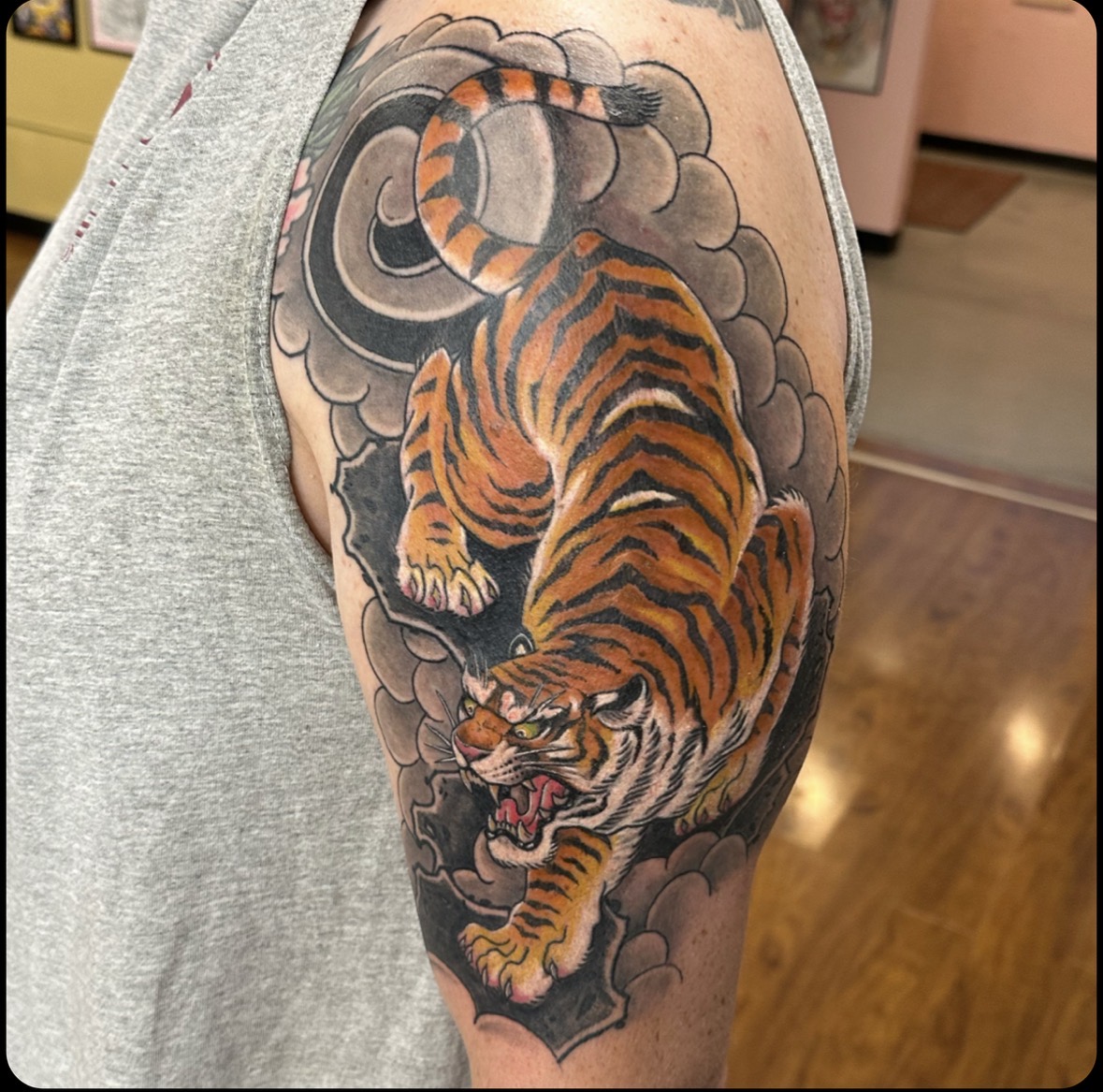 Tiger tattoo on upper arm with clouds and smoke, detailed orange and black stripes, fierce expression, and open mouth.