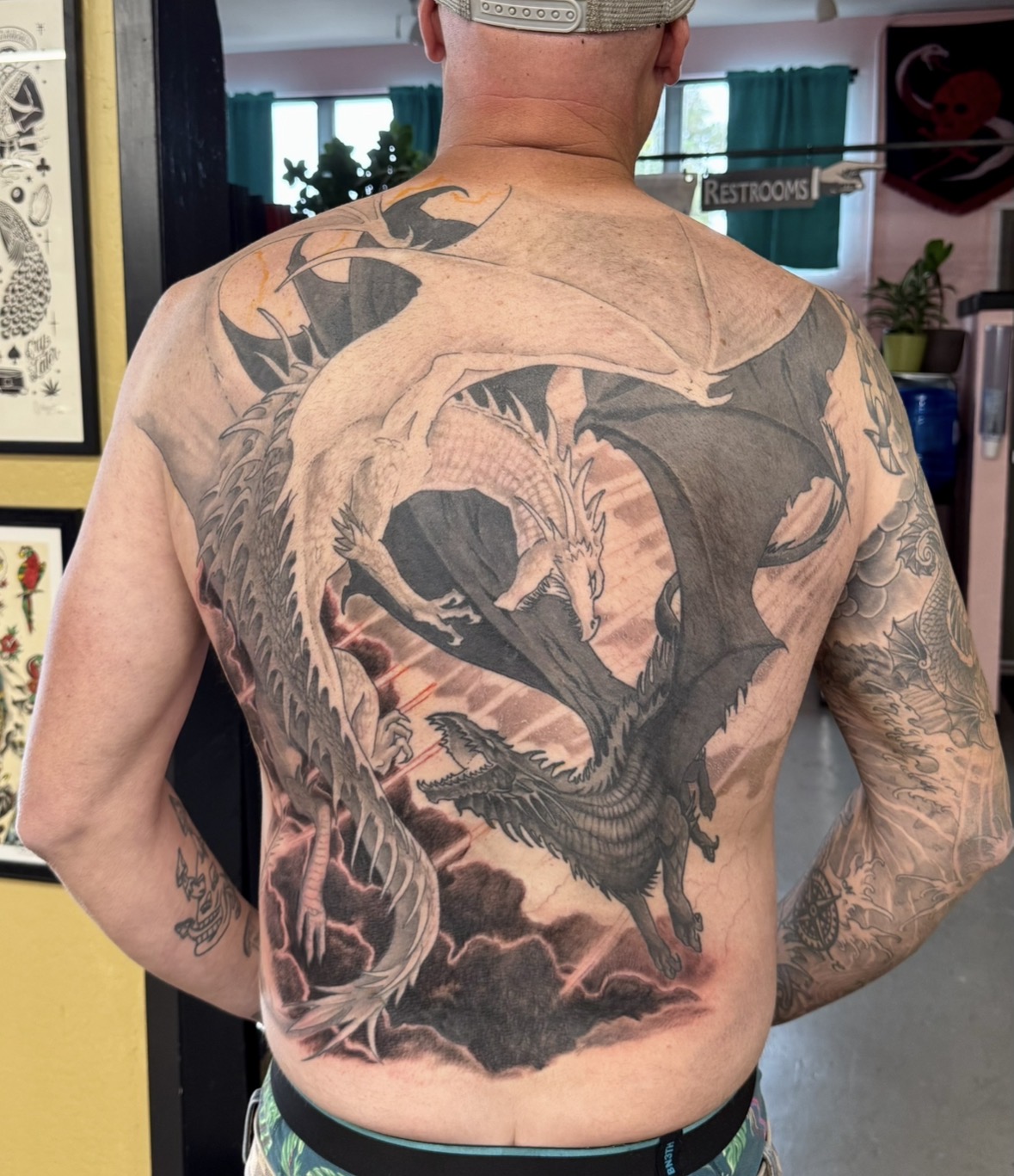 Back view of a person with a large dragon tattoo covering the back and shoulders, detailed black and gray design.