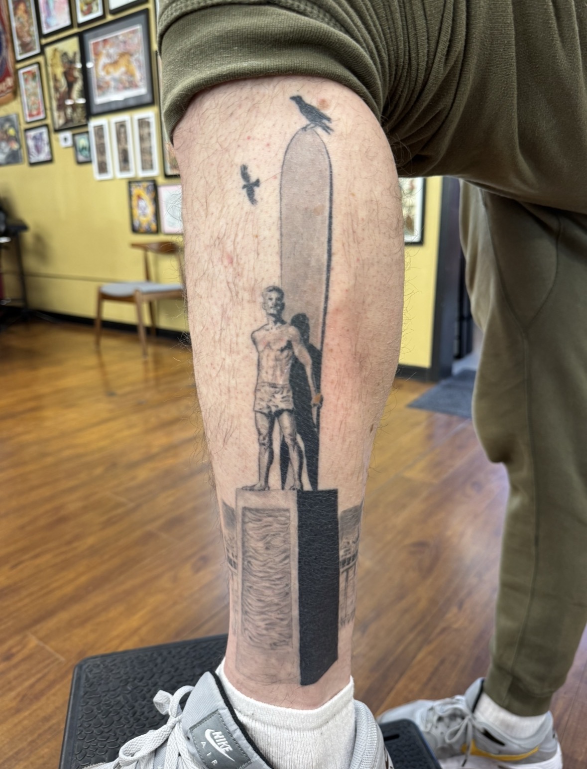 Tattoo of a person standing on a block, with a tall vertical shape and birds flying above, on a person's arm.