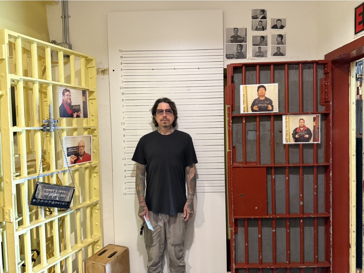 Man with glasses and tattoos stands between yellow and red metal structures with photos on them, in an indoor space.