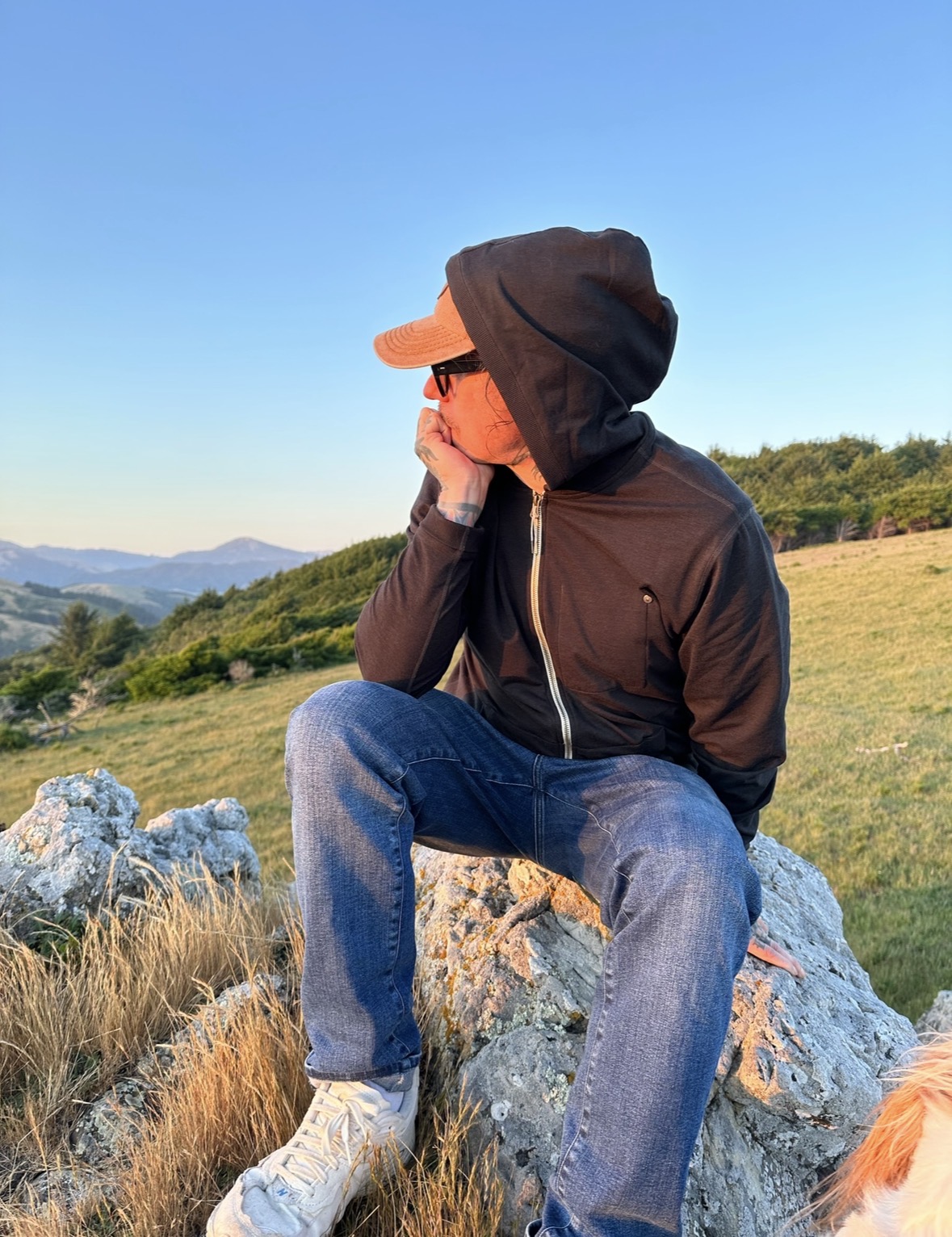 Person sitting outdoors on rocks, wearing a hoodie, cap, and sunglasses, with a landscape of hills and sky in background.