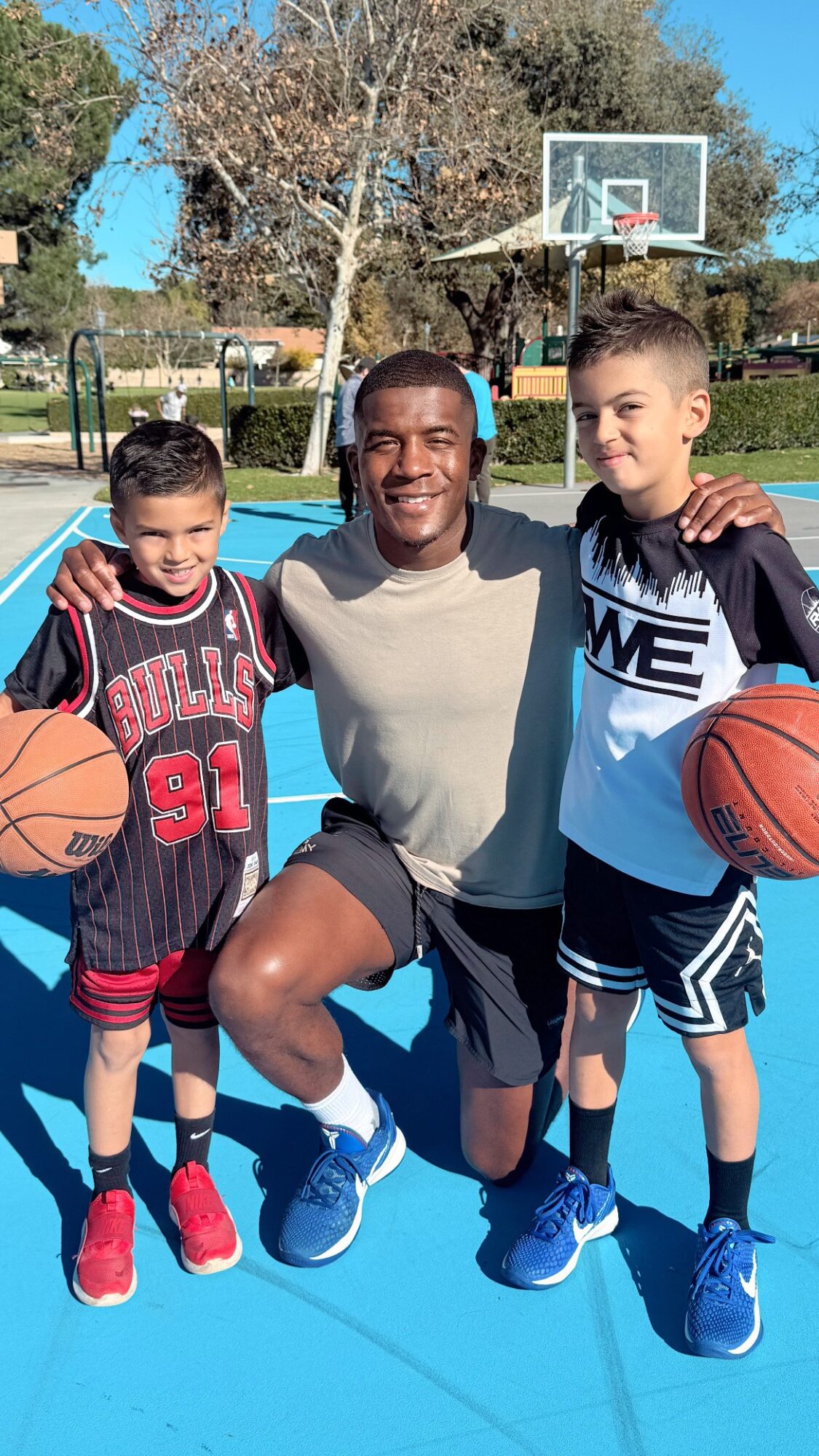 Three people on a basketball court, two children holding basketballs, adult in the middle, smiling, outdoor setting.