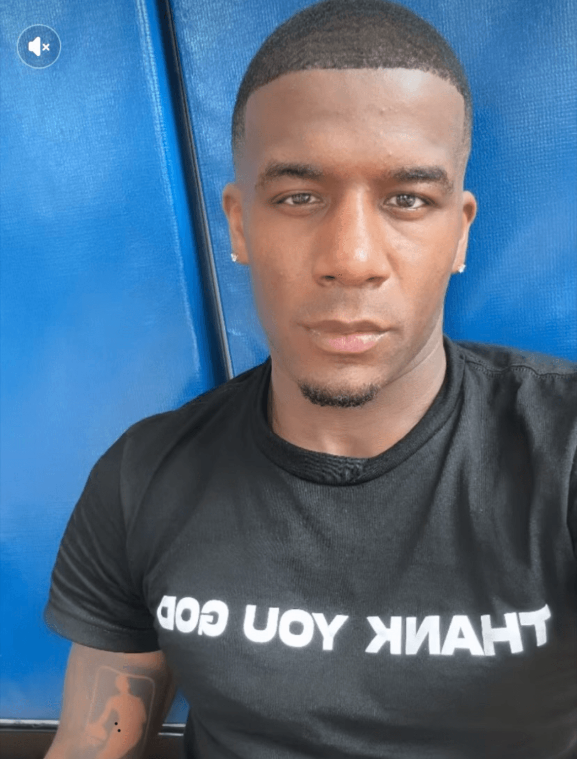 Young man with short hair and earrings, wearing a black T-shirt with white text, standing against a blue background.