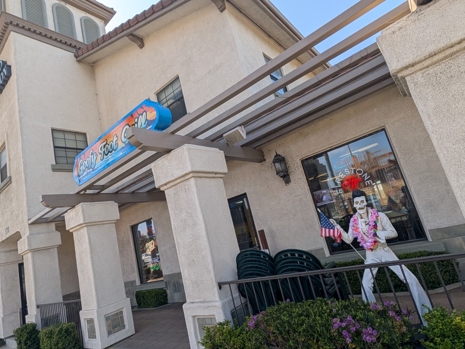 Building exterior with a colorful sign, large windows, and a mannequin dressed as a clown holding American flags, surrounded by plants.