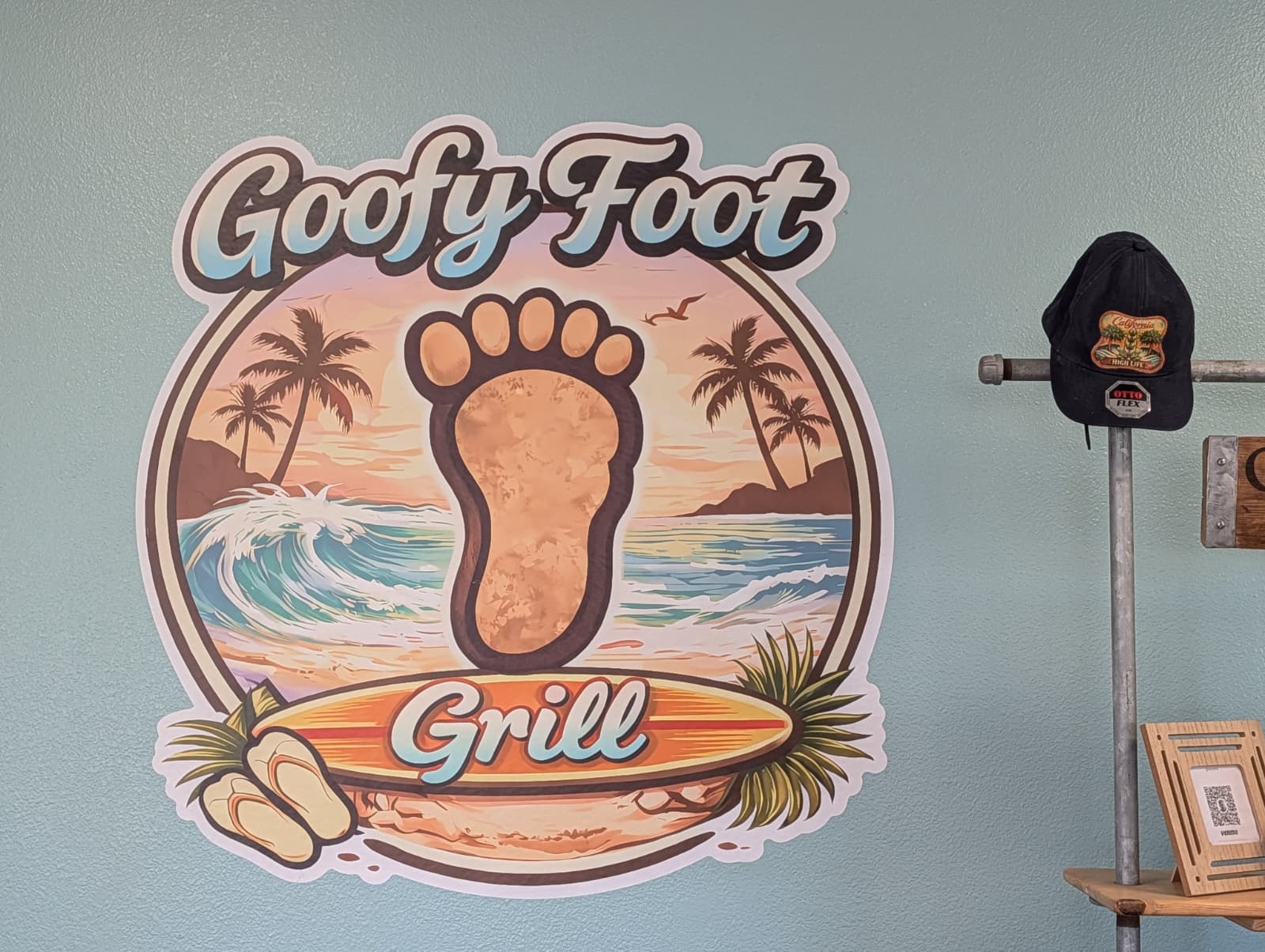 Wall sign with a large footprint, palm trees, ocean waves, and sunset, reading 'Goofy Foot Grill'.