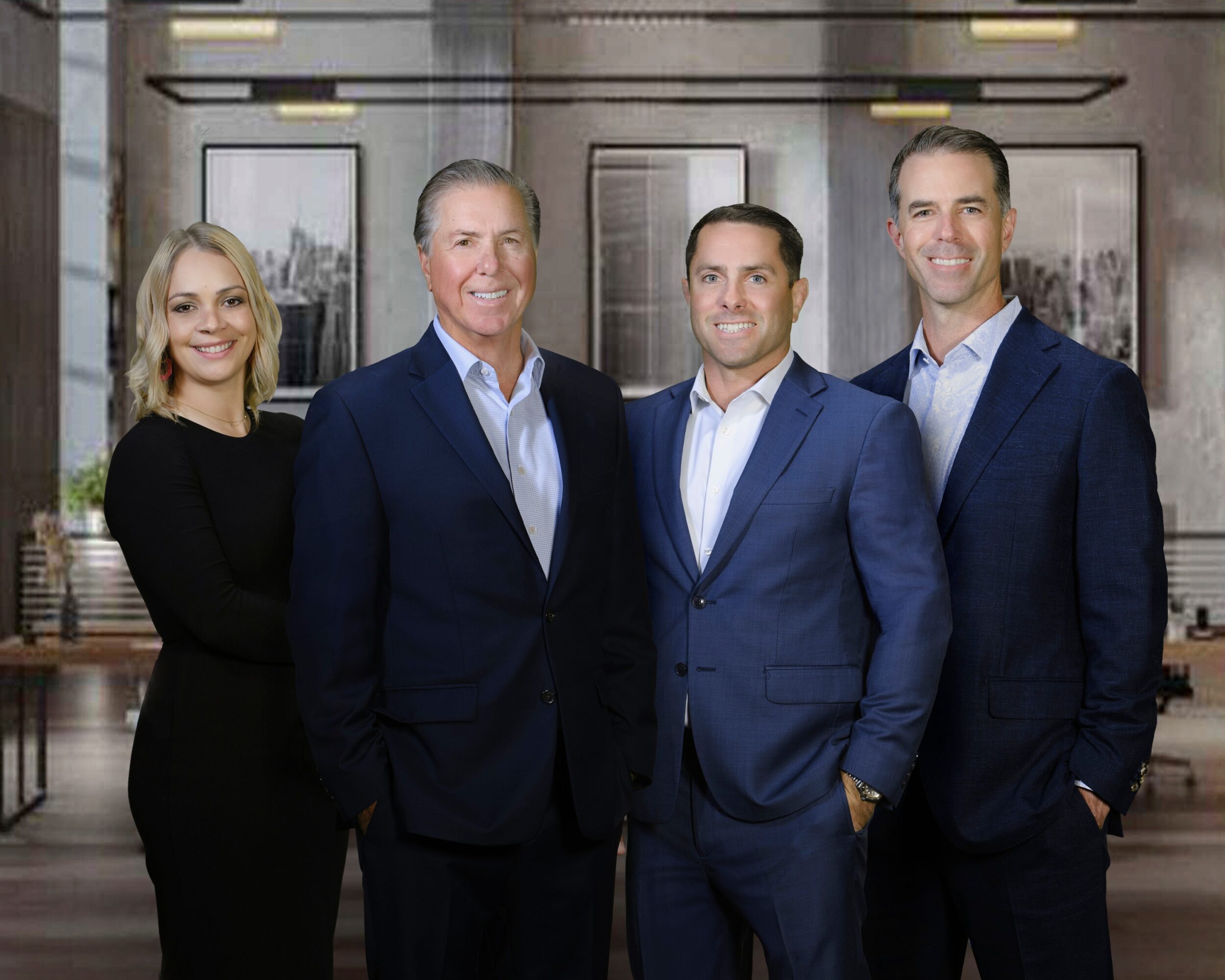 Four professionally dressed people standing in a modern office with large windows and cityscape view behind them.