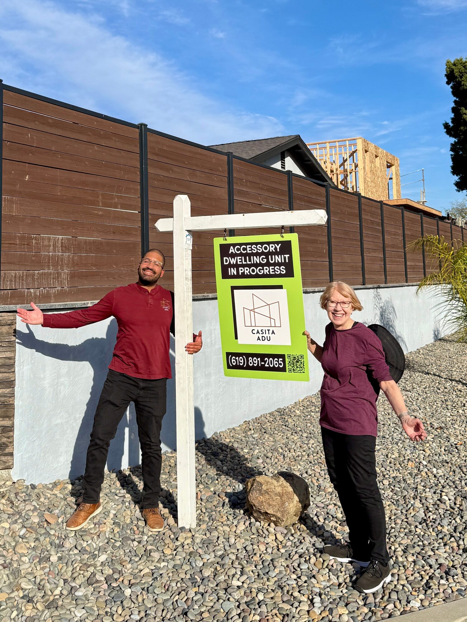 Two people standing outdoors near a sign, with a wooden fence and construction site in background, on gravel ground.