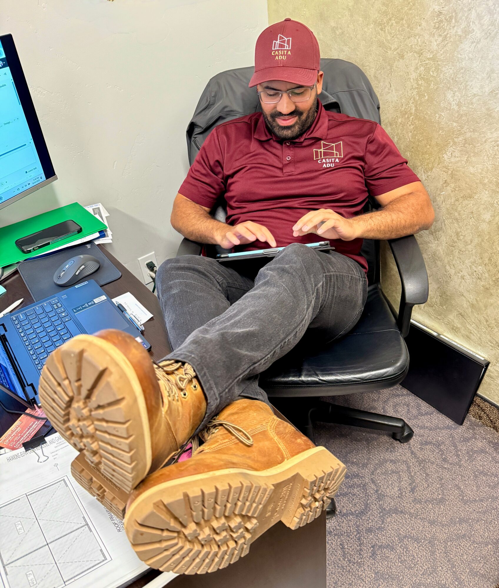 Man sitting in an office chair with legs crossed, wearing a red cap and shirt, using a tablet, surrounded by office supplies.