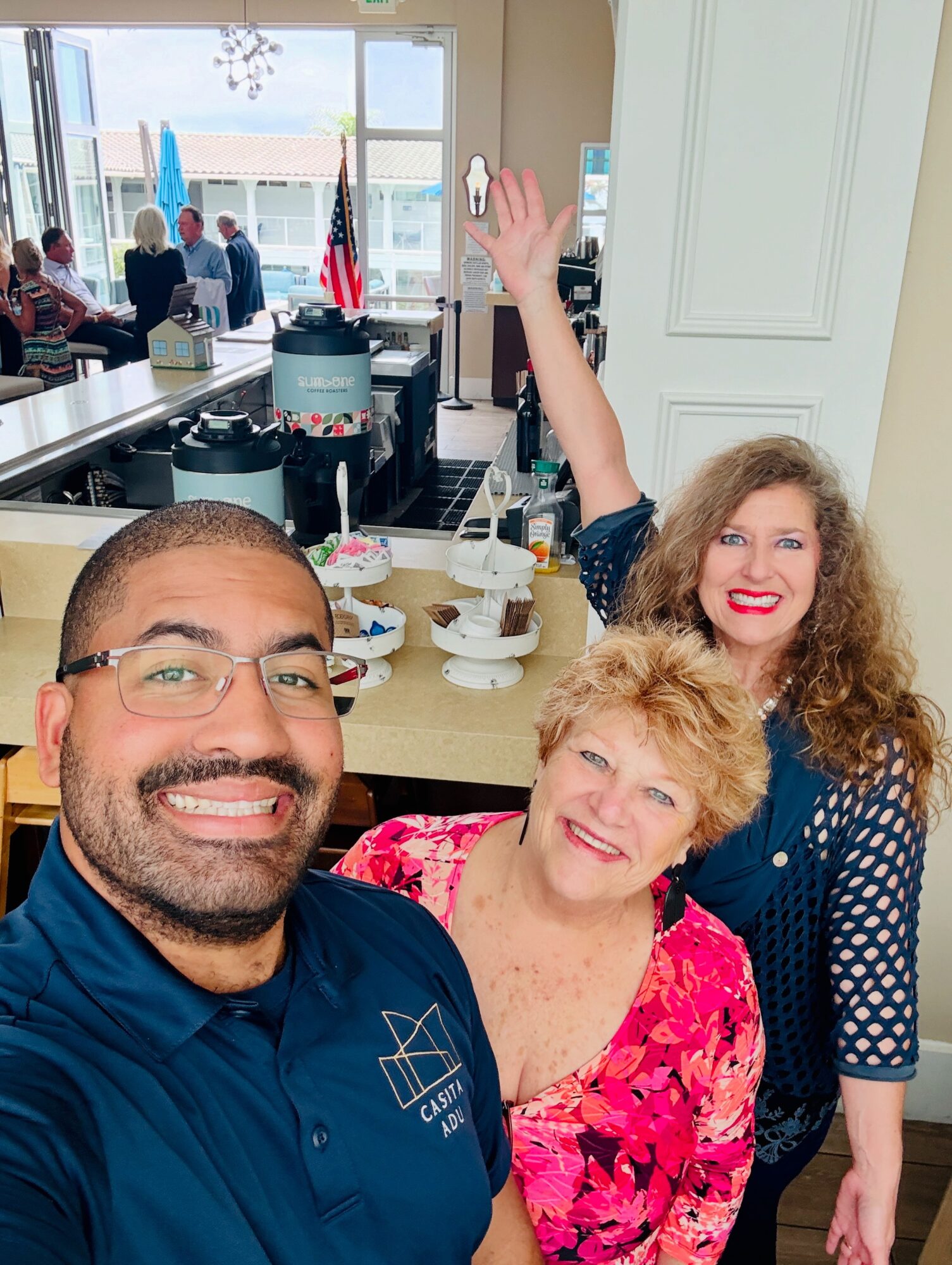 Three people smiling and taking a selfie in a bright room with large windows and a view outside, with a table and food in the background.