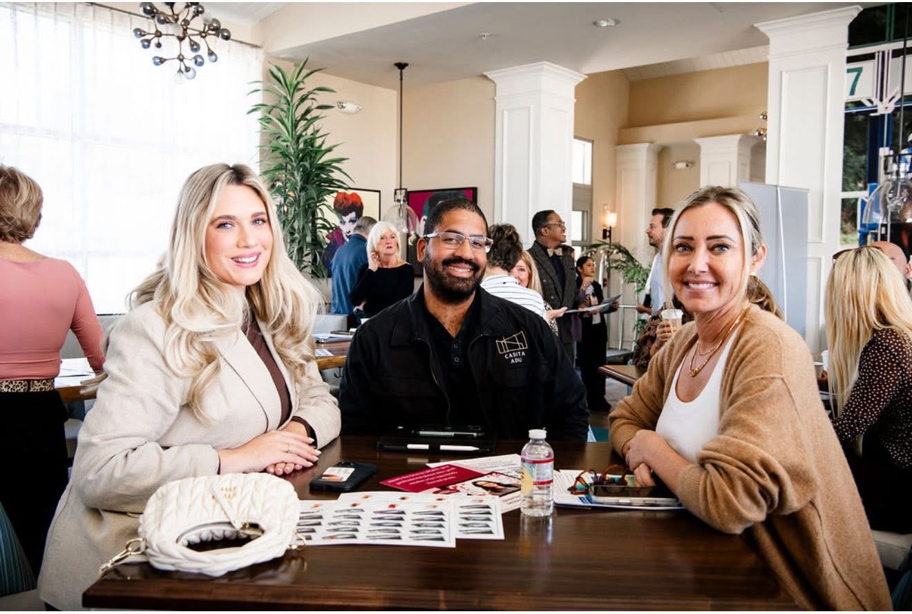 Three people sit at a table in a busy indoor setting, smiling at the camera, with a plant and other people in the background.