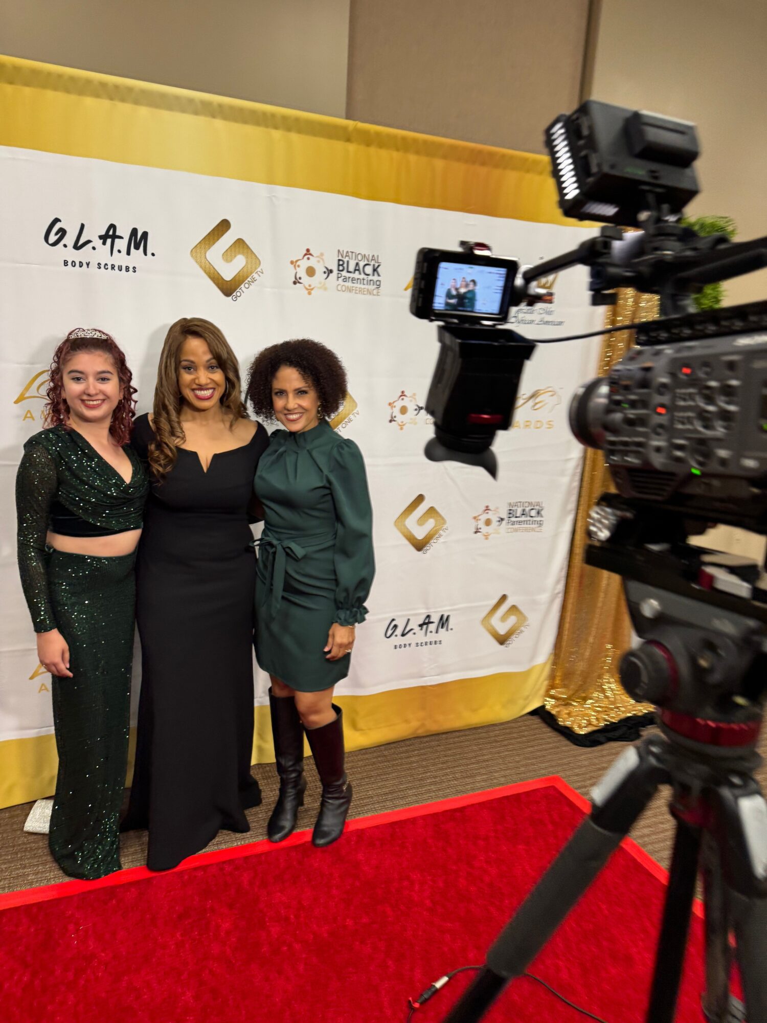 Three women standing on a red carpet in front of a white backdrop with logos, surrounded by cameras and equipment.