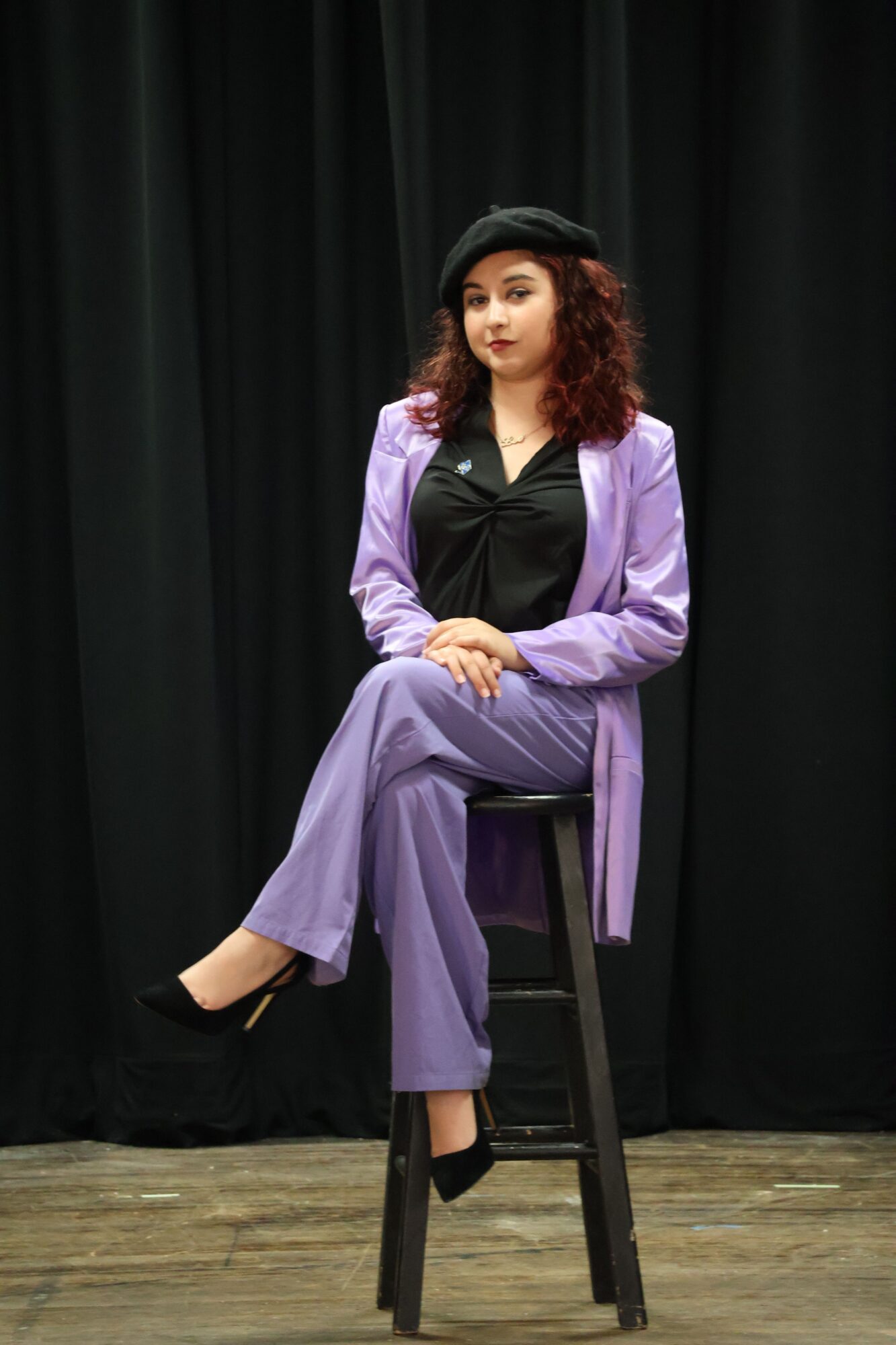 Woman with curly hair in purple pants and black top sitting on a chair against a black curtain background.