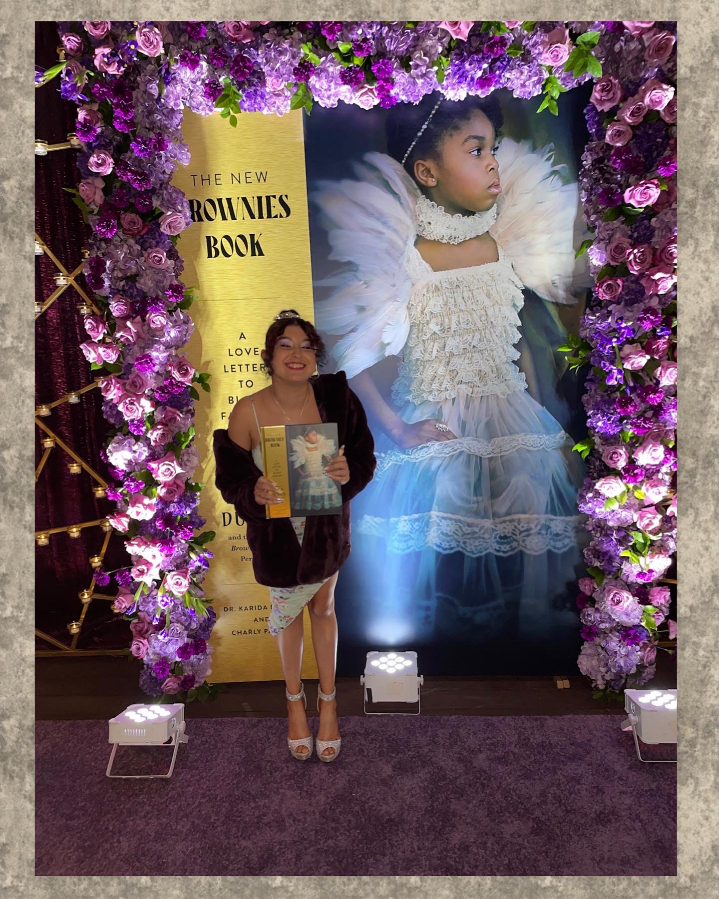 Woman standing in front of a large portrait of a girl in a white dress, surrounded by purple flowers and a yellow sign.