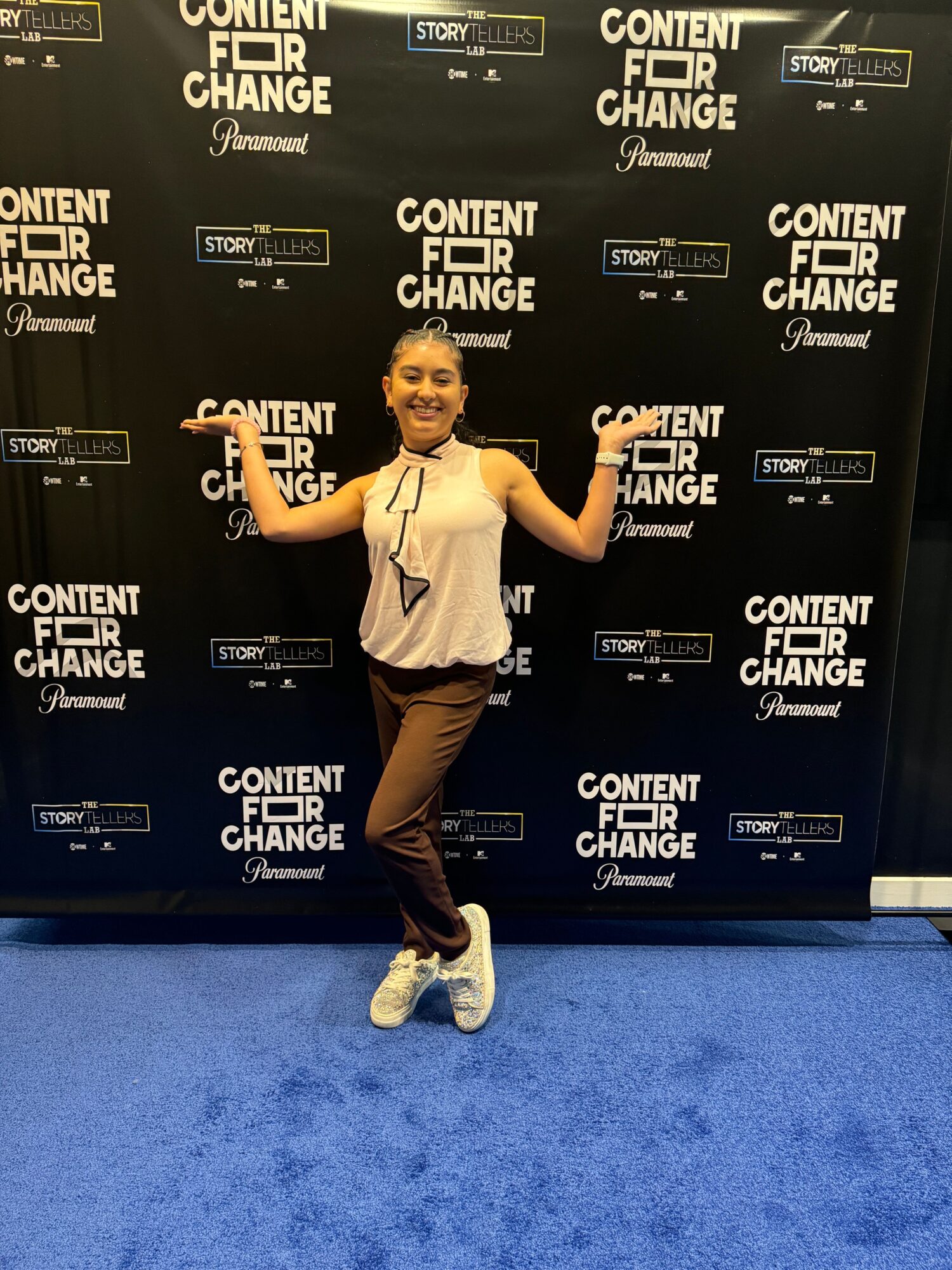 Person standing in front of a black backdrop with white text, wearing a white shirt, brown pants, and sneakers, posing with one arm raised.
