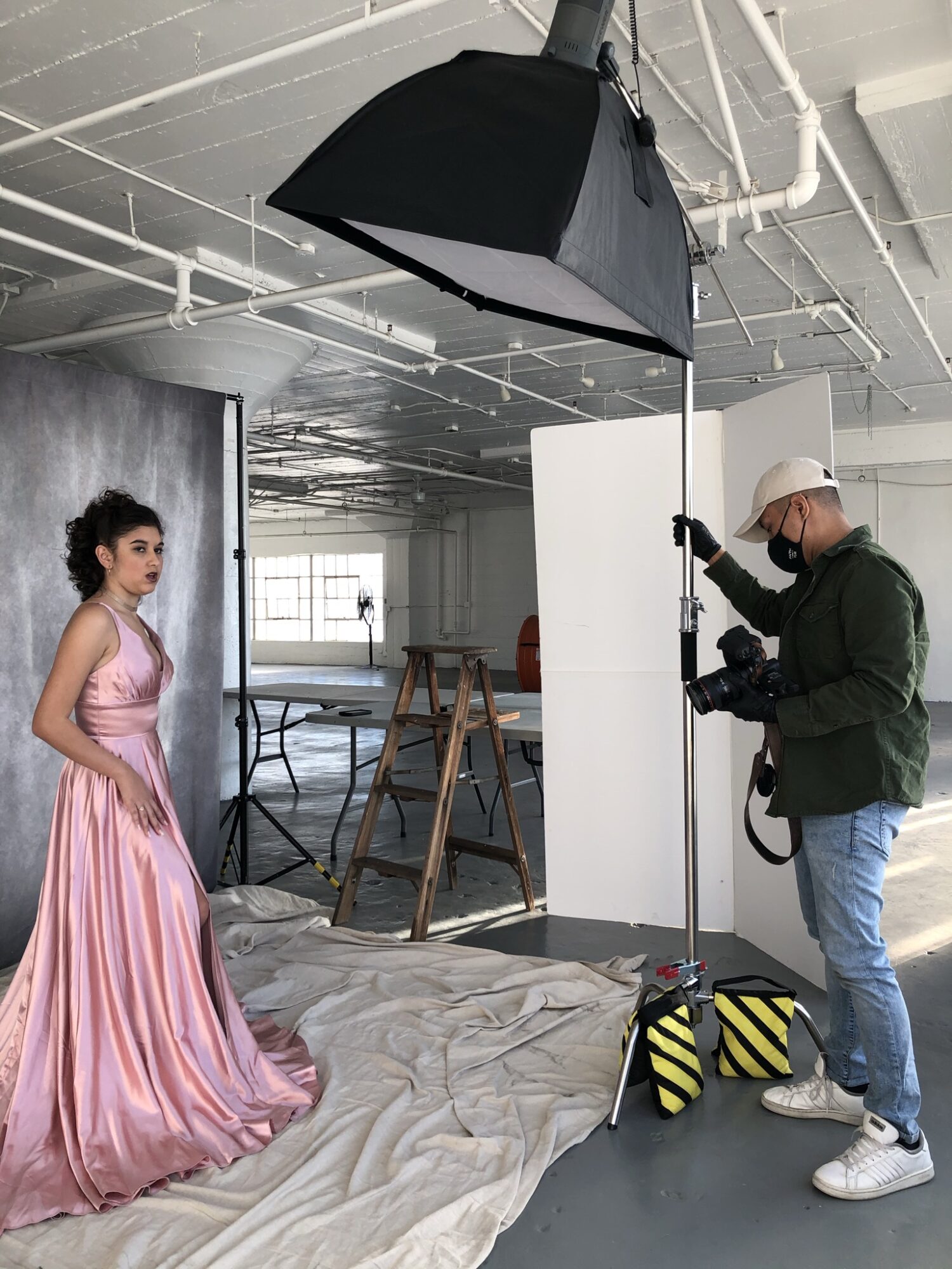 A woman in a pink dress stands in a photography studio with a photographer adjusting equipment, large softbox overhead, and a white backdrop.