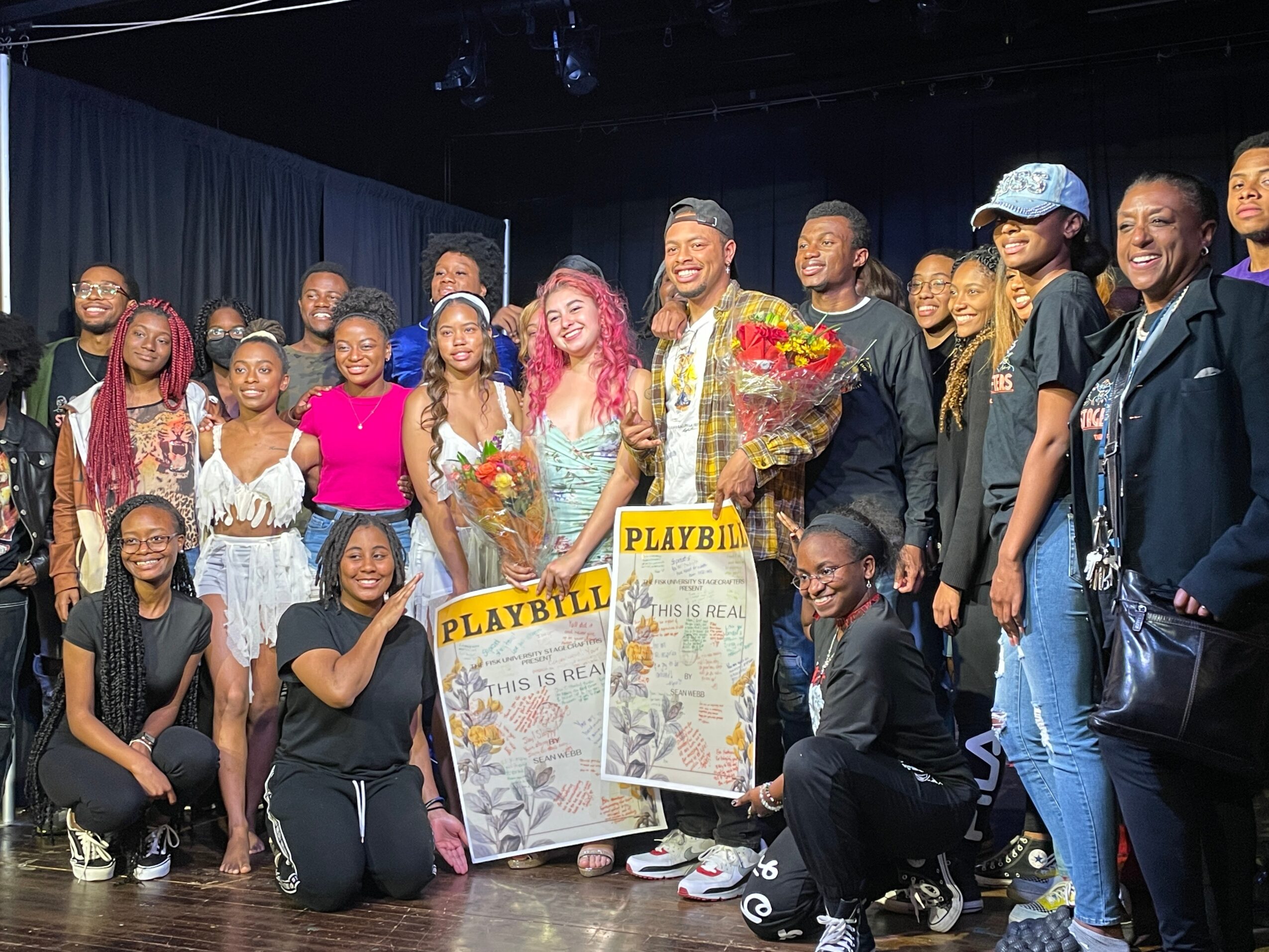 Group of people on stage celebrating, some holding Playbill posters, smiling, with a dark curtain background.