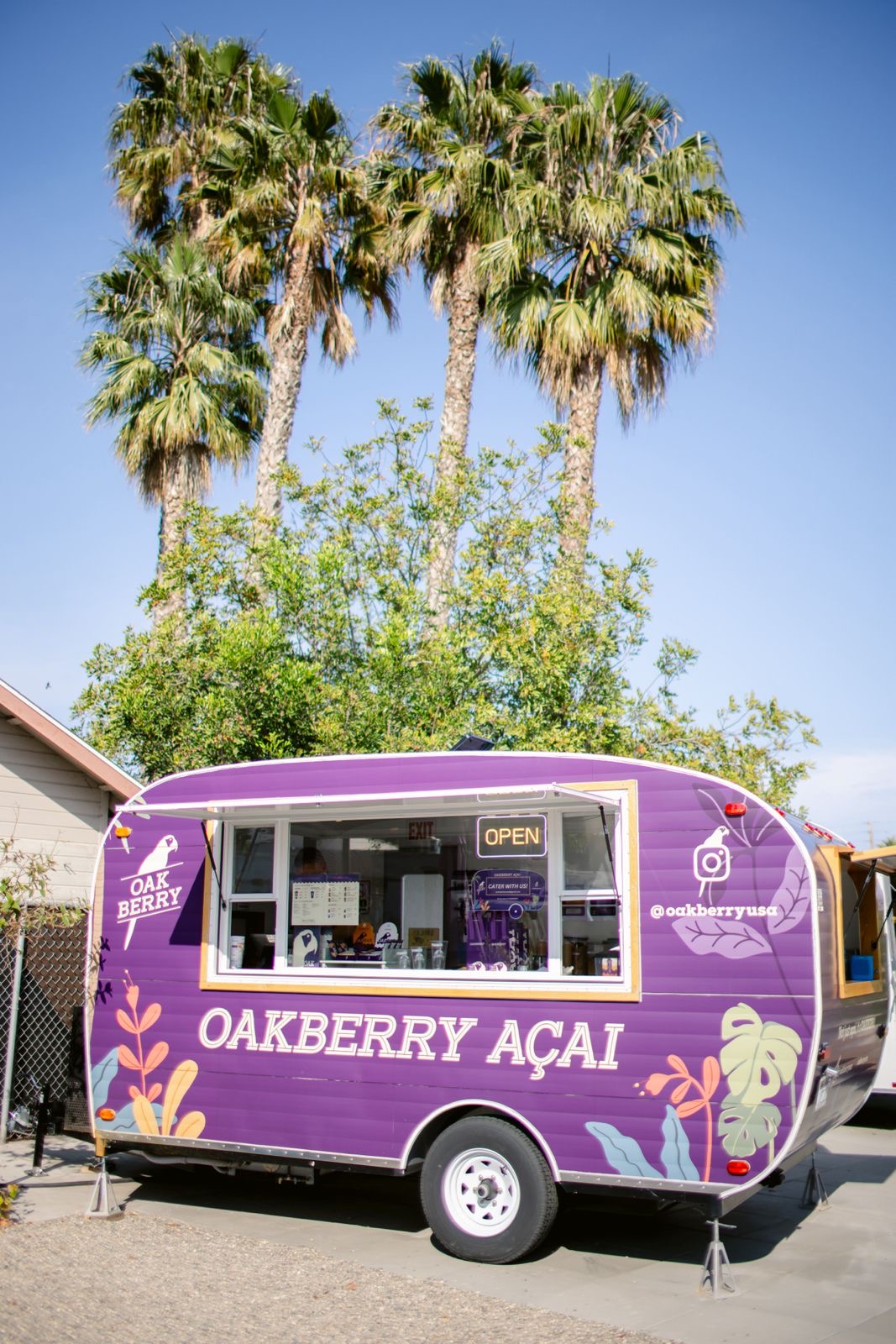 Purple food truck with white text and floral design, parked in front of tall palm trees and a small building, sunny day.