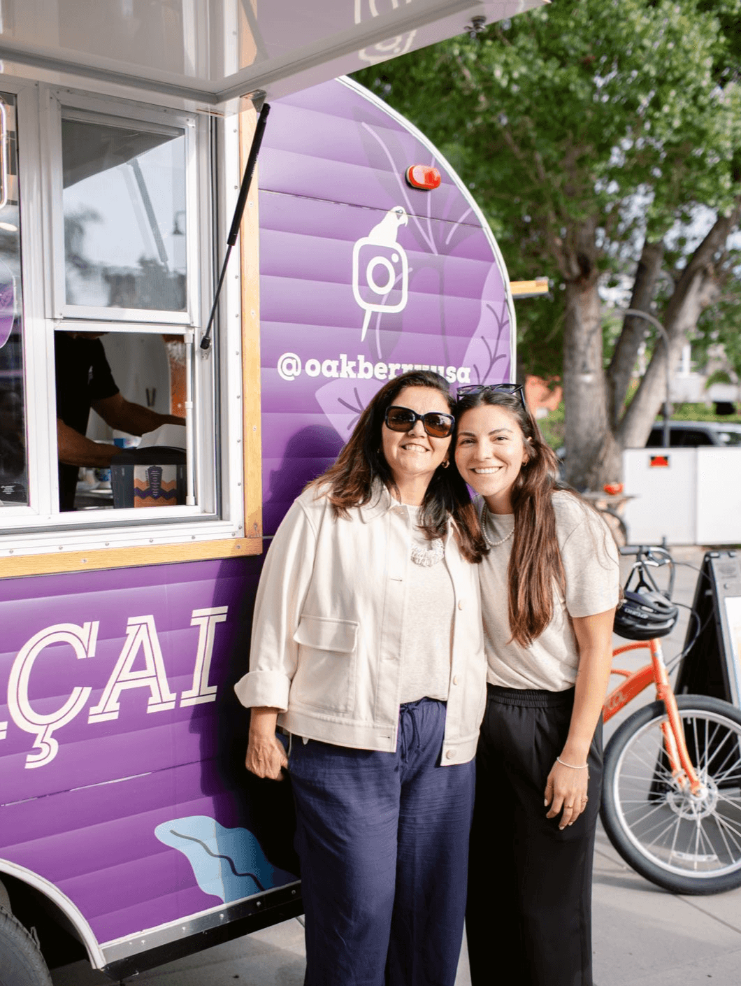 Two women stand outside a purple food truck with social media icons and text, smiling at the camera.
