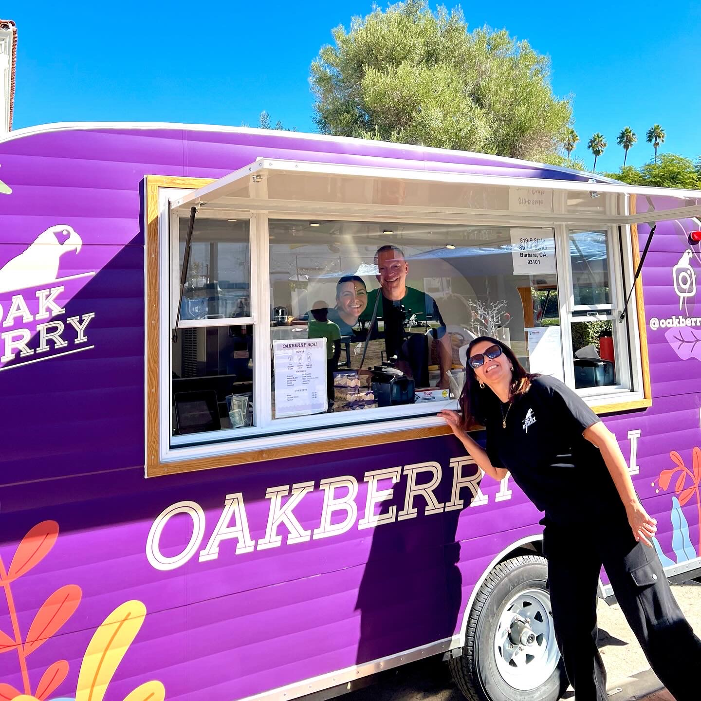 Person leaning on purple food truck with window, two people inside, bright blue sky, trees, and palm trees in background.