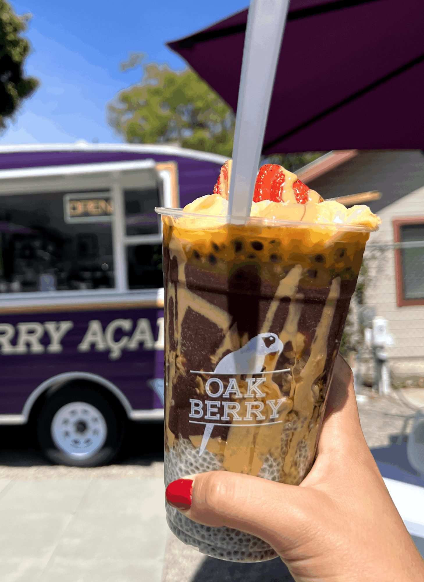 Hand holding a passion fruit and berry smoothie with a straw and fruit topping, outdoors on a sunny day.