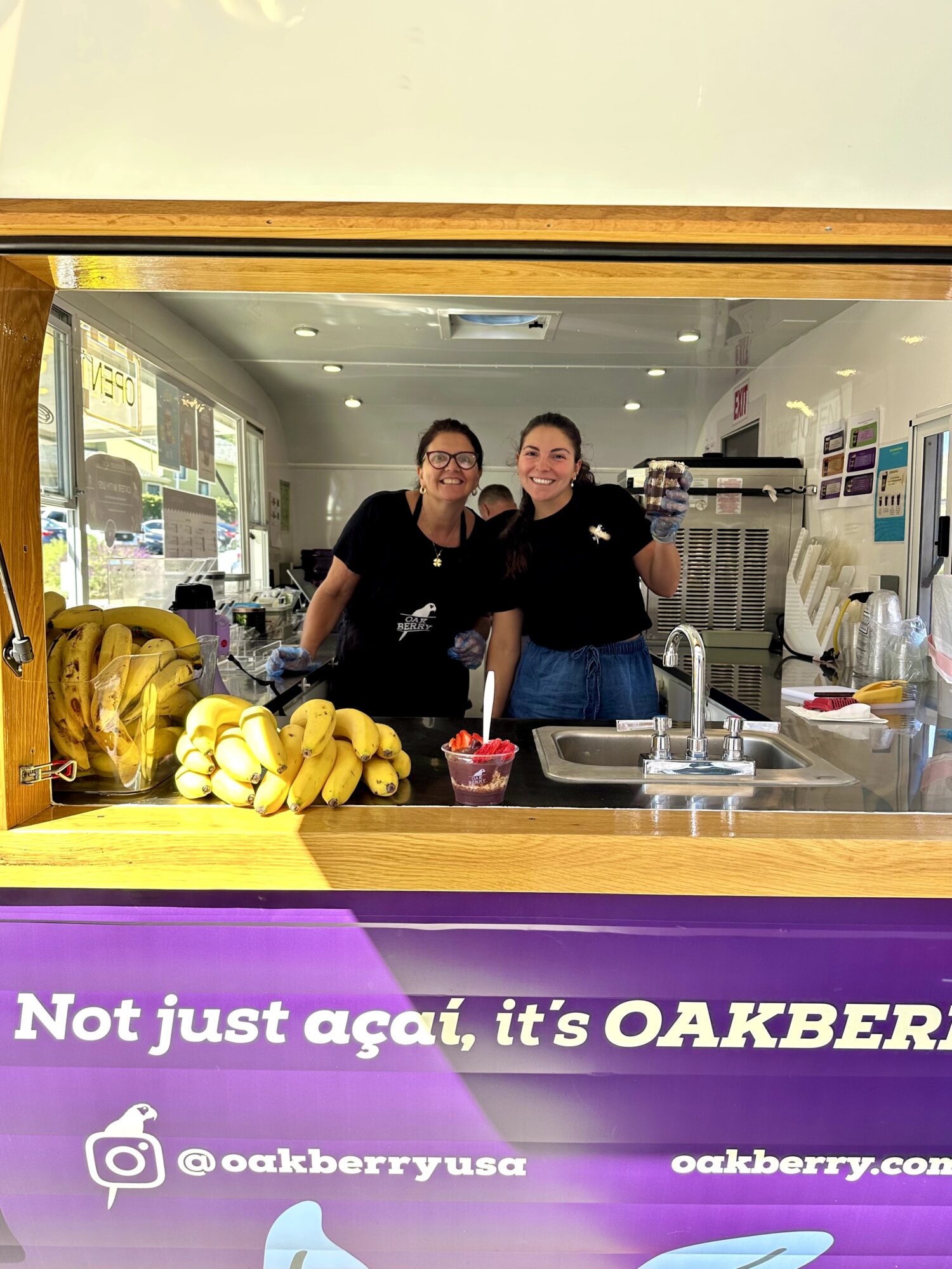 Two women smiling and taking a selfie in a kitchen, with bananas and kitchen appliances visible behind them.