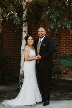 A bride and groom stand outdoors in front of a brick wall and trees, smiling and holding hands.