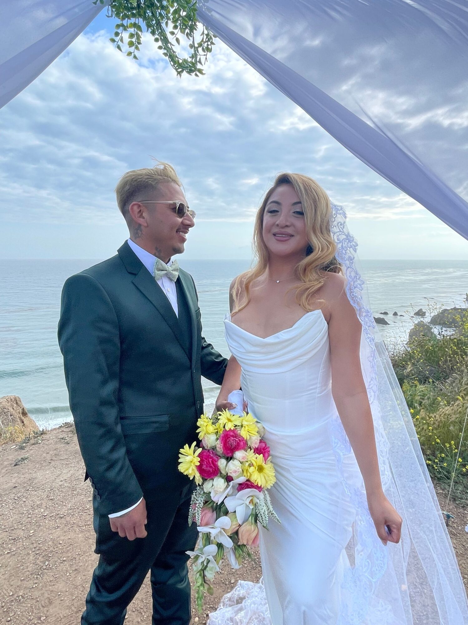 Bride and groom standing outdoors near water, bride in white dress, groom in dark suit, holding a bouquet of flowers.