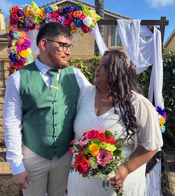 A couple stands outdoors in front of a floral arch, holding a bouquet, with a house and blue sky in background.